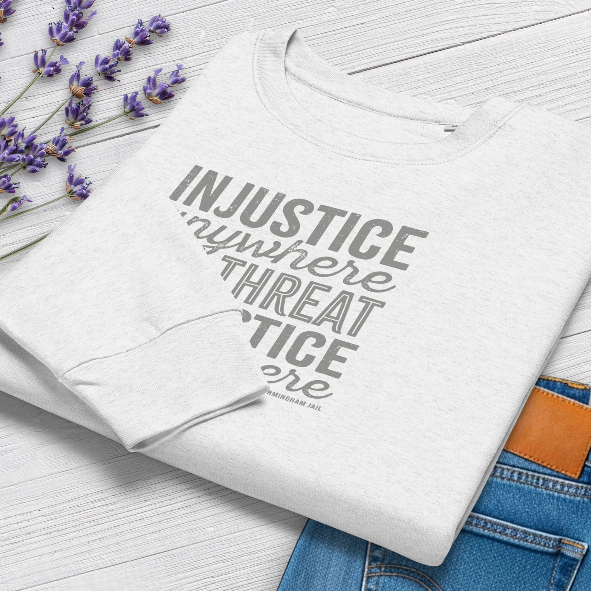 Folded grey sweatshirt with bold text reading “Injustice anywhere is a threat to justice everywhere” quote from Martin Luther King Jr, MLK Jr, Letters from Birmingham jail — civil rights protest apparel for activists. Styled with jeans and flowers.