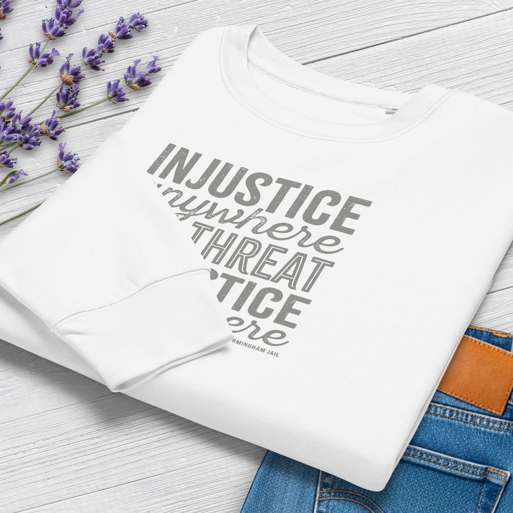 Folded white sweatshirt with bold text reading “Injustice anywhere is a threat to justice everywhere” quote from Martin Luther King Jr, MLK Jr, Letters from Birmingham jail — civil rights protest apparel for activists. Styled with jeans and flowers.