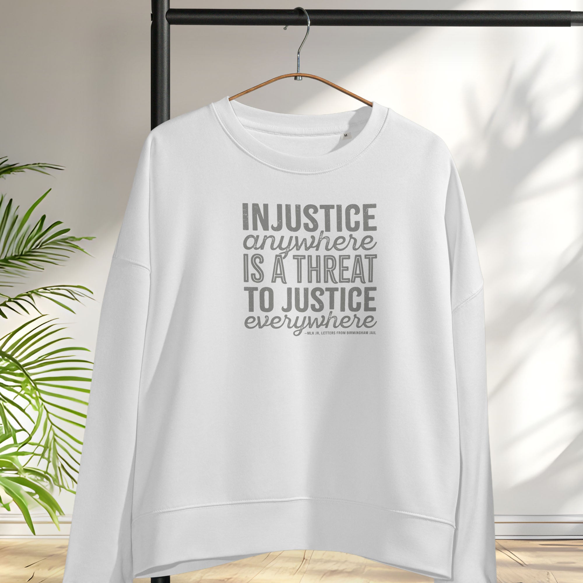 White sweatshirt with bold text reading “Injustice anywhere is a threat to justice everywhere” quote from Martin Luther King Jr, MLK Jr, Letters from Birmingham jail — civil rights protest apparel for activists. Hanging on a hanger.