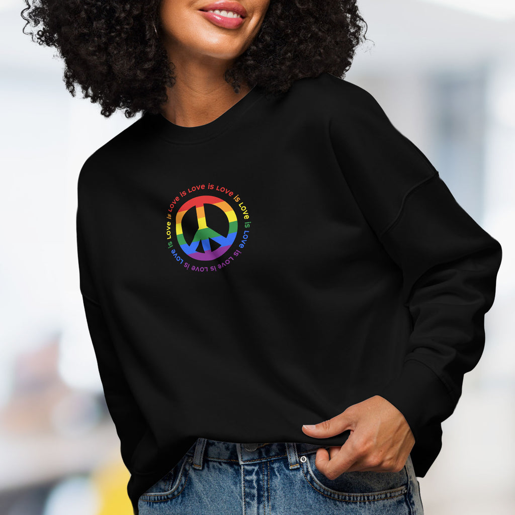 Female model wearing black sweatshirt with LGBTQ pride graphic — a peace sign in the LGBTQ flag colors, with the text "love is love" repeated in a circle around the peace sign.