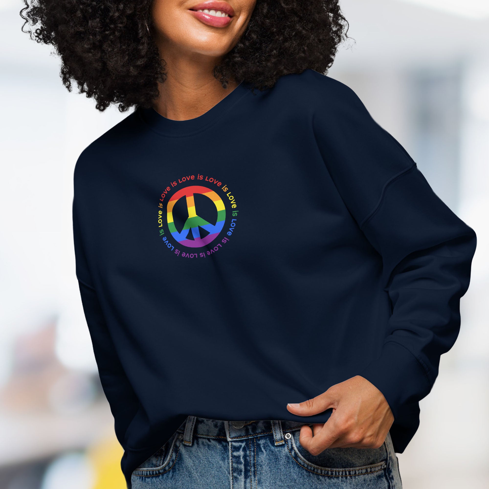 Female model wearing navy sweatshirt with LGBTQ pride graphic — a peace sign in the LGBTQ flag colors, with the text "love is love" repeated in a circle around the peace sign.