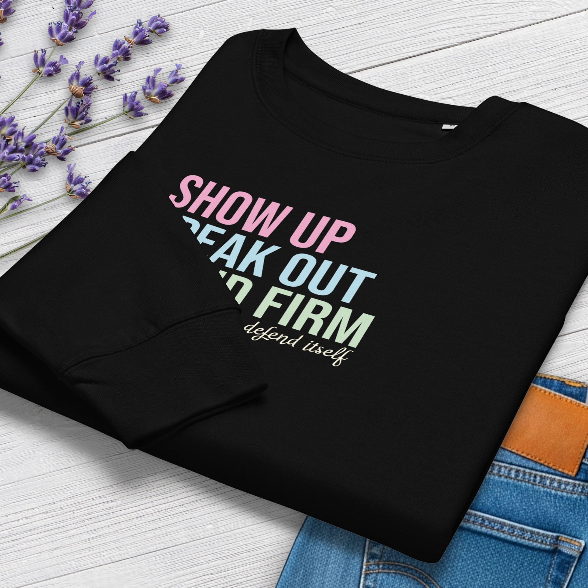 Folded black women's sweatshirt with bright pink, blue and green bold text that says "show up, speak out, stand firm" and subtle script text below that reads "democracy doesn't defend itself." Styled with a pair of jeans and flowers.