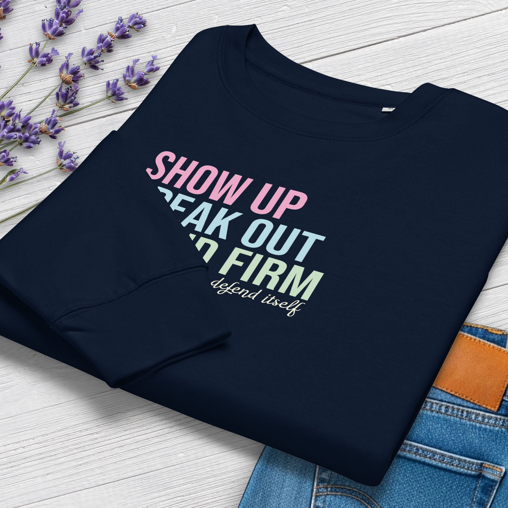 Folded navy women's sweatshirt with bright pink, blue and green bold text that says "show up, speak out, stand firm" and subtle script text below that reads "democracy doesn't defend itself." Styled with a pair of jeans and flowers.