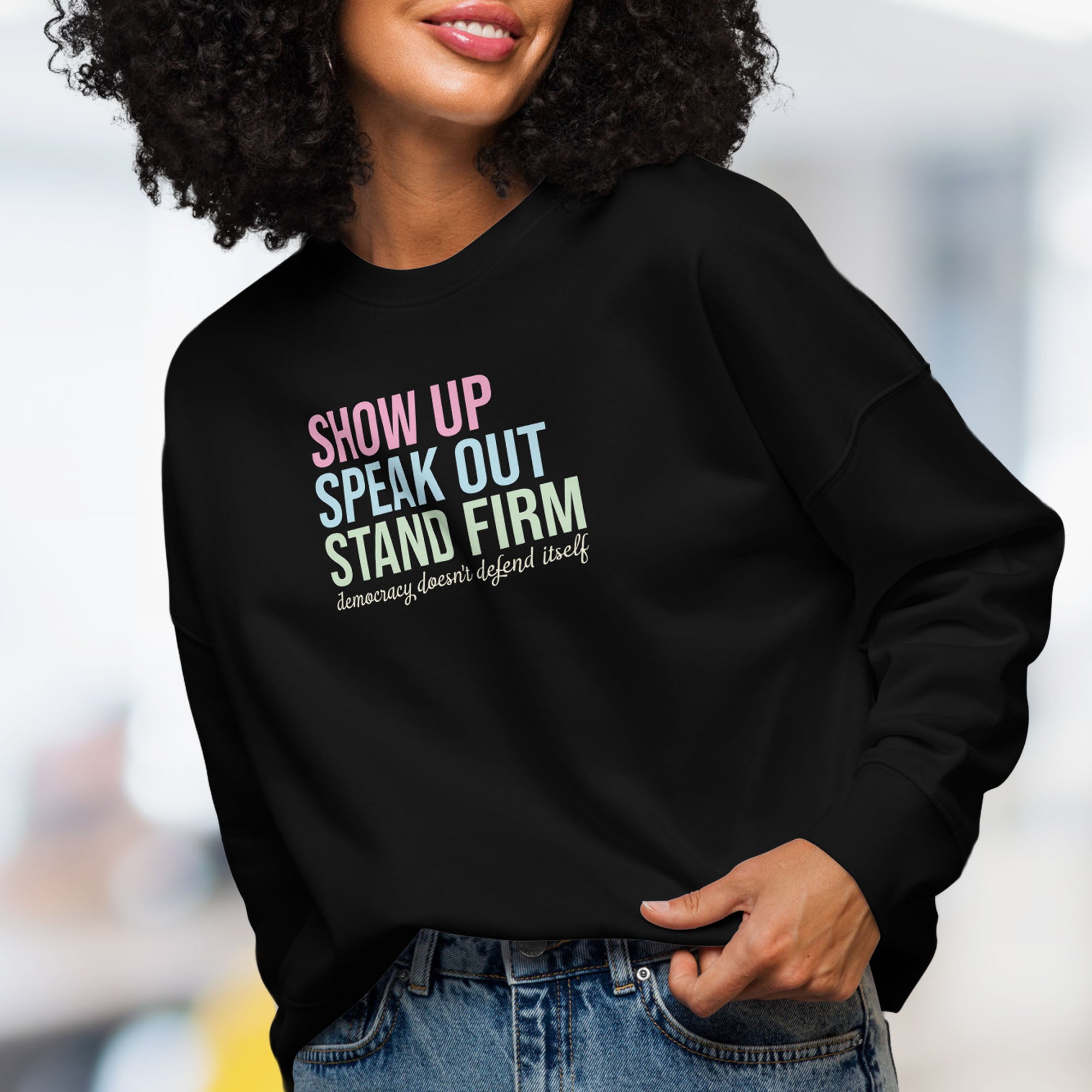 Female model wearing women's black sweatshirt with bright pink, blue and green bold text that says "show up, speak out, stand firm" and subtle script text below that reads "democracy doesn't defend itself."