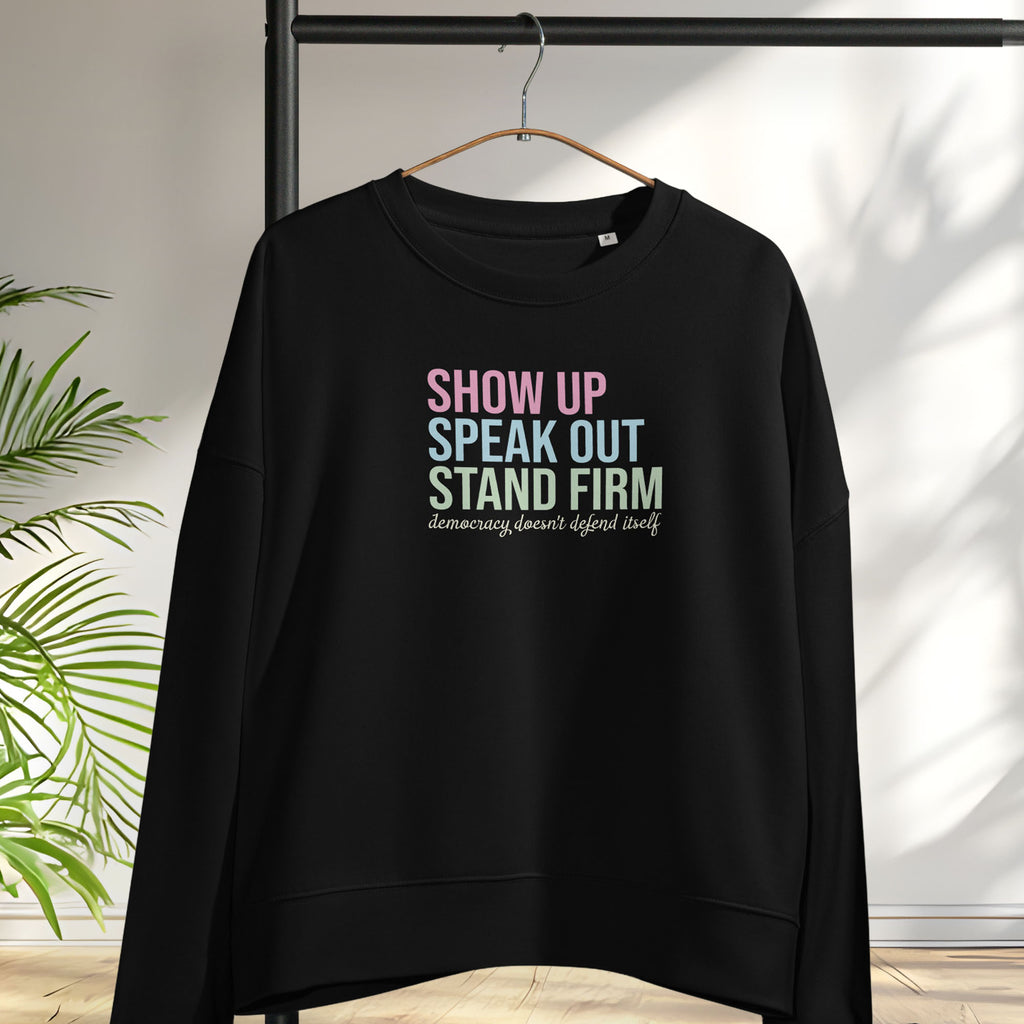 Black women's sweatshirt with bright pink, blue and green bold text that says "show up, speak out, stand firm" and subtle script text below that reads "democracy doesn't defend itself." Hanging on a hanger.