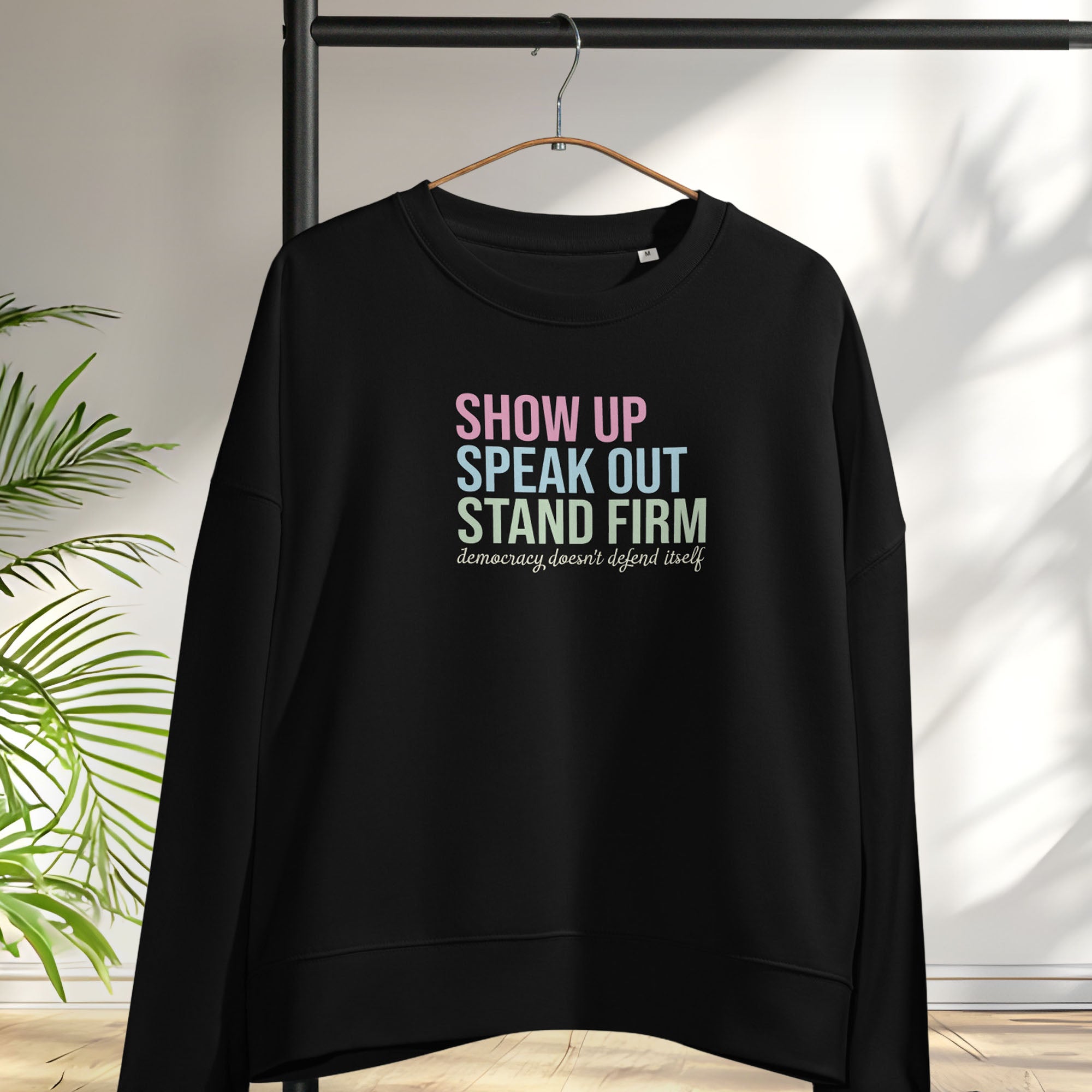 Black women's sweatshirt with bright pink, blue and green bold text that says "show up, speak out, stand firm" and subtle script text below that reads "democracy doesn't defend itself." Hanging on a hanger.