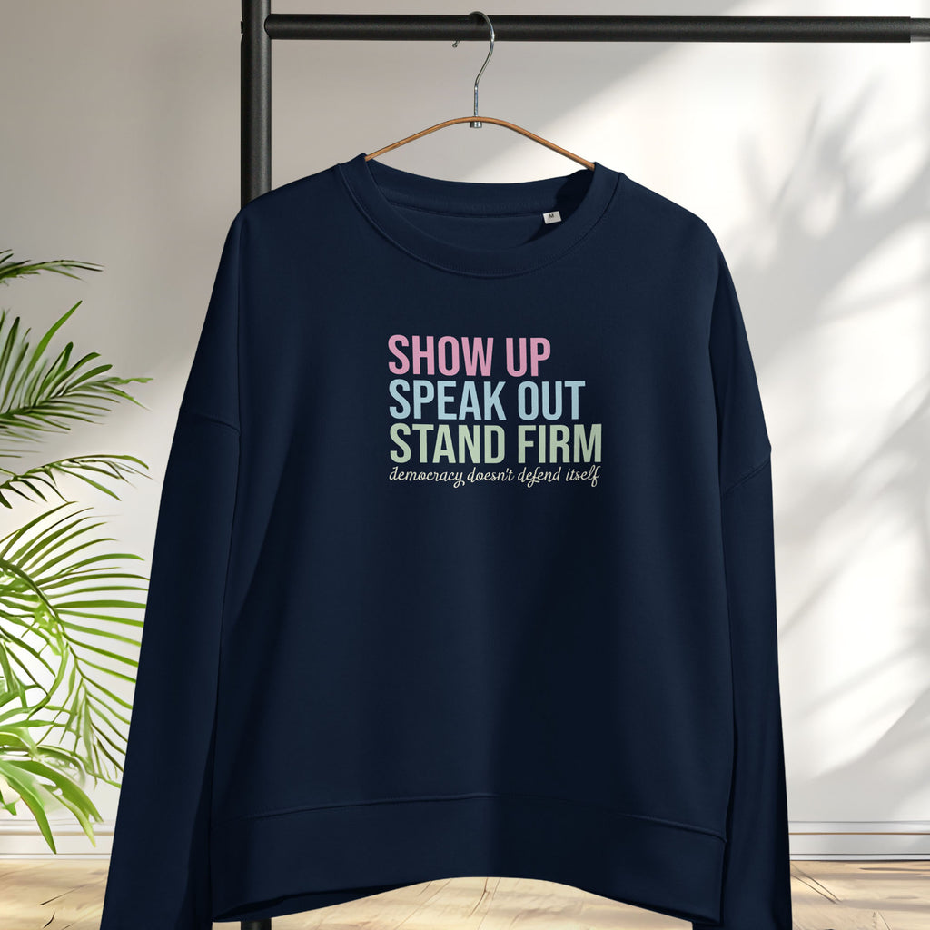 Black women's sweatshirt with bright pink, blue and green bold text that says "show up, speak out, stand firm" and subtle script text below that reads "democracy doesn't defend itself." Hanging on a hanger.