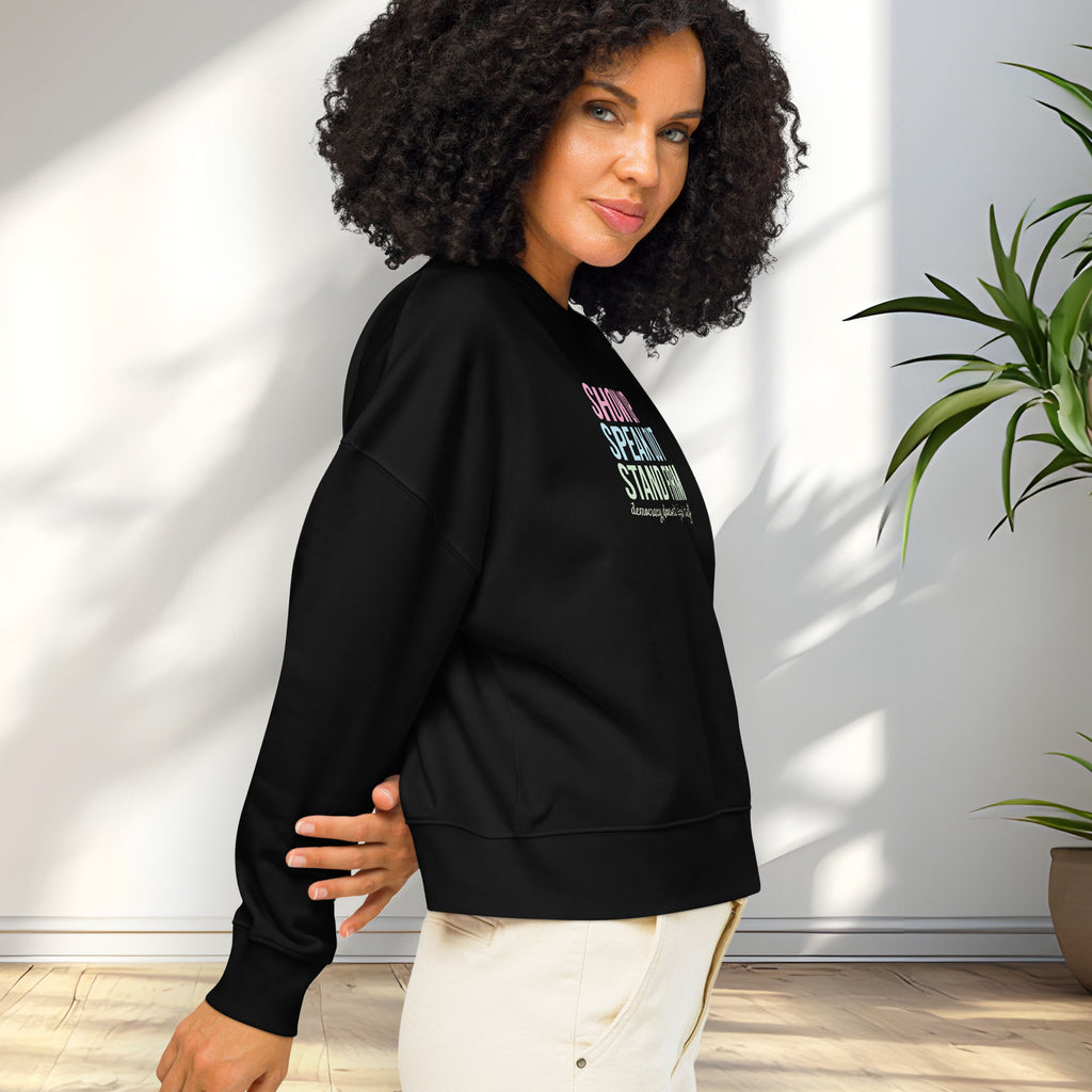 Side view of female model wearing black women's sweatshirt with bright pink, blue and green bold text that says "show up, speak out, stand firm" and subtle script text below that reads "democracy doesn't defend itself."