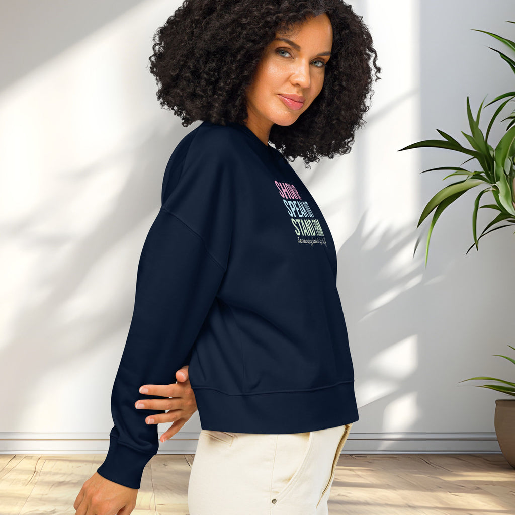 Side view of female model wearing black women's sweatshirt with bright pink, blue and green bold text that says "show up, speak out, stand firm" and subtle script text below that reads "democracy doesn't defend itself."