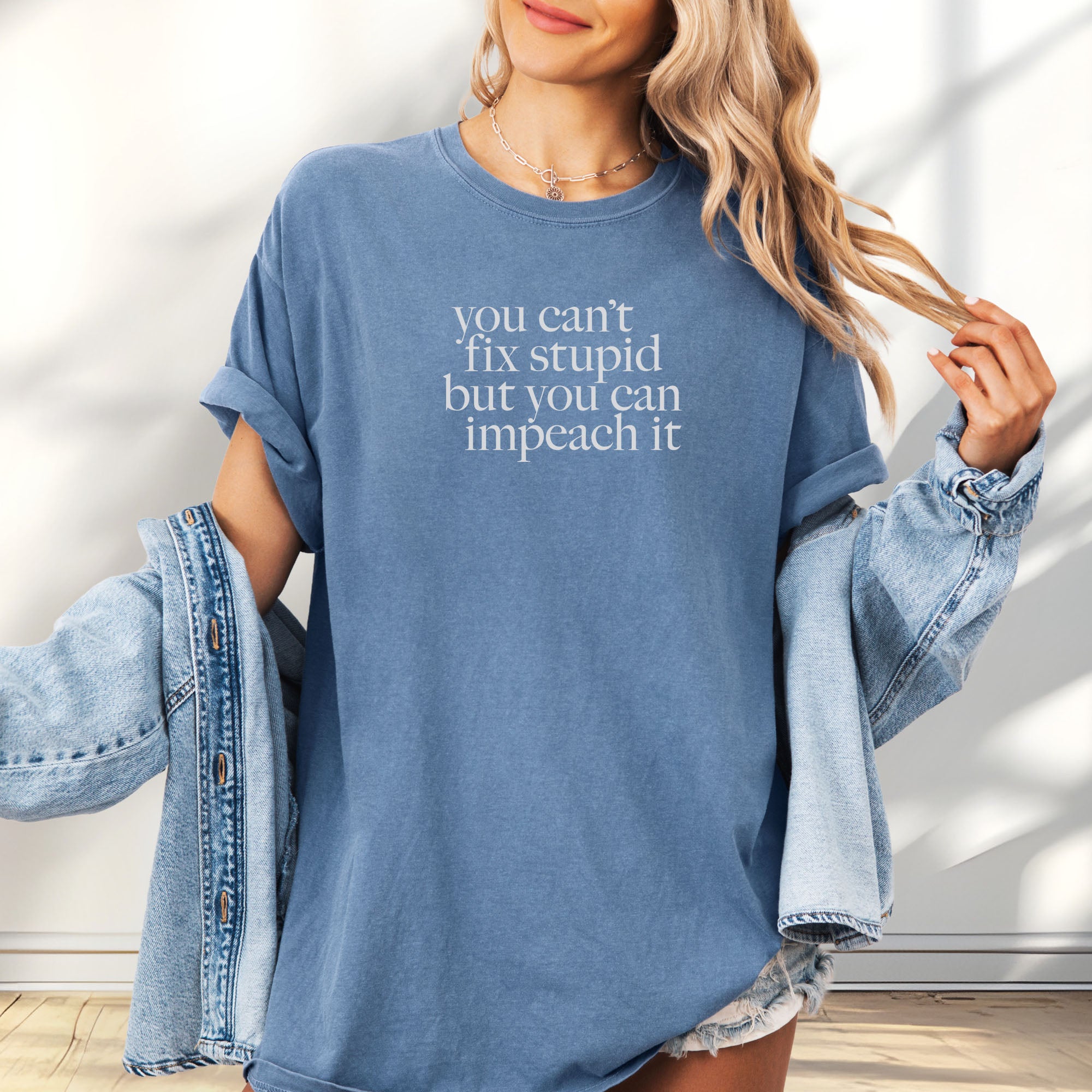 Female model wearing oversized blue jean political t-shirt with serif text reading “You can’t fix stupid but you can impeach it.".