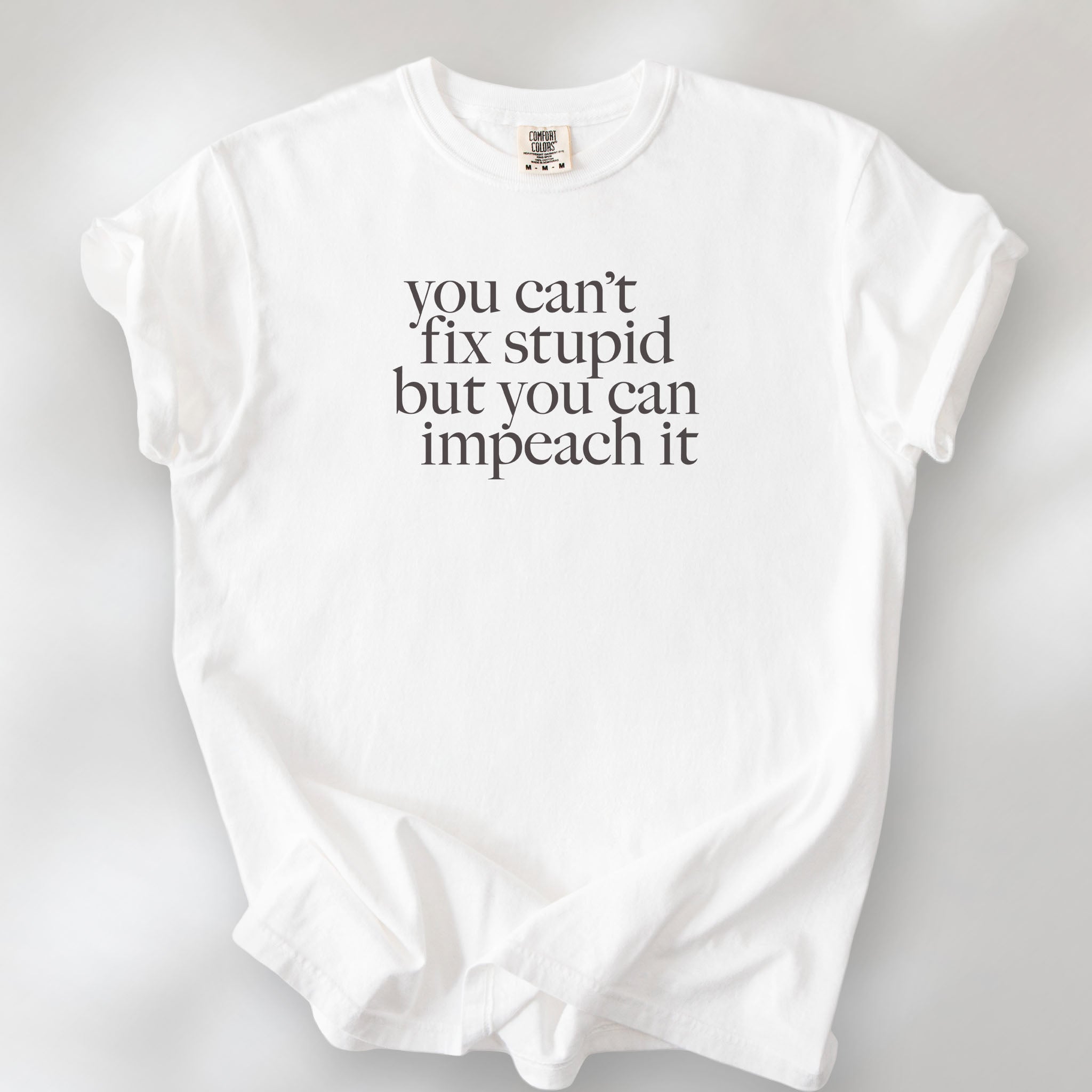 White political t-shirt with serif text reading “You can’t fix stupid but you can impeach it.".
