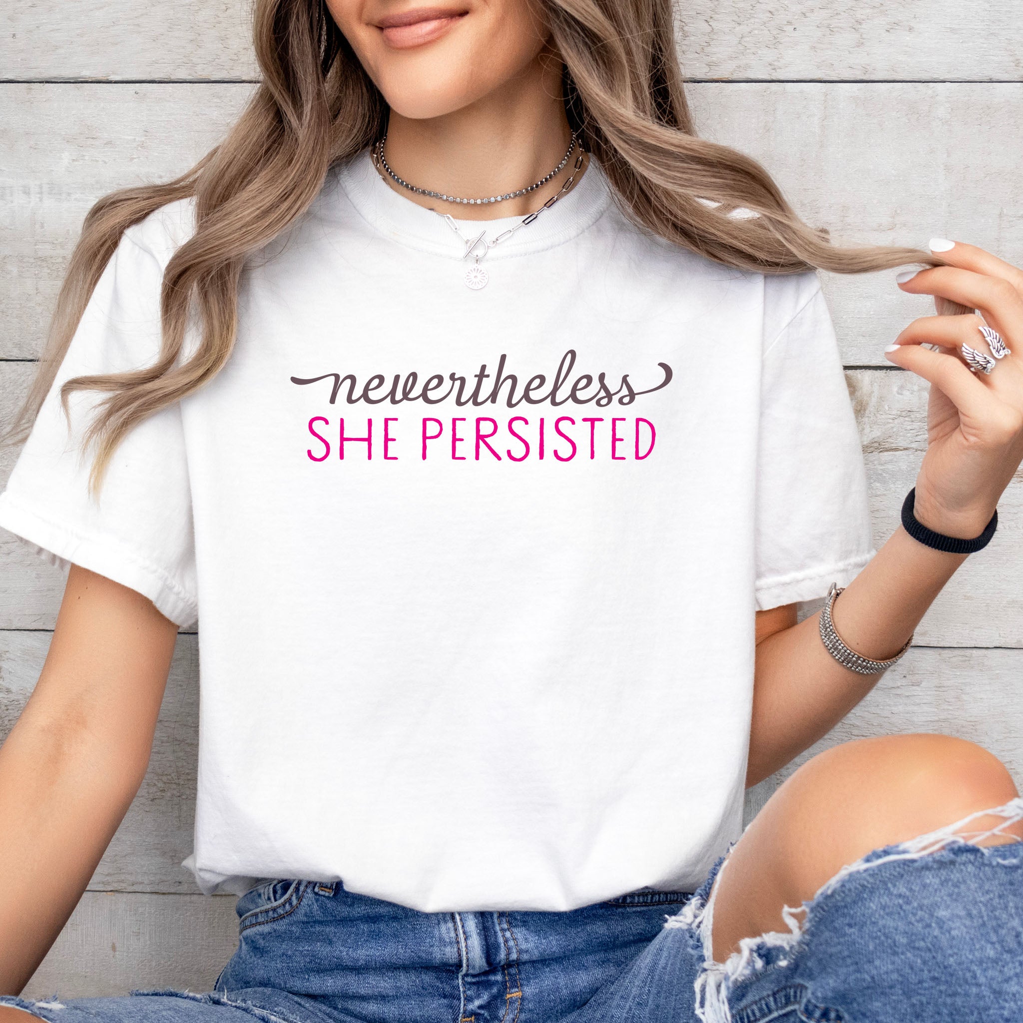 Female model wearing white Comfort Colors garment-dyed tee with stylized text graphic that reads "nevertheless she persisted."