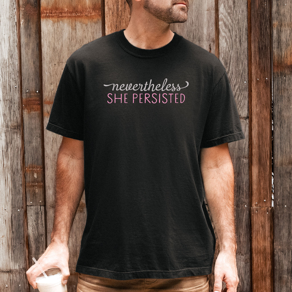 Male model wearing black Comfort Colors garment-dyed tee with stylized text graphic that reads "nevertheless she persisted."