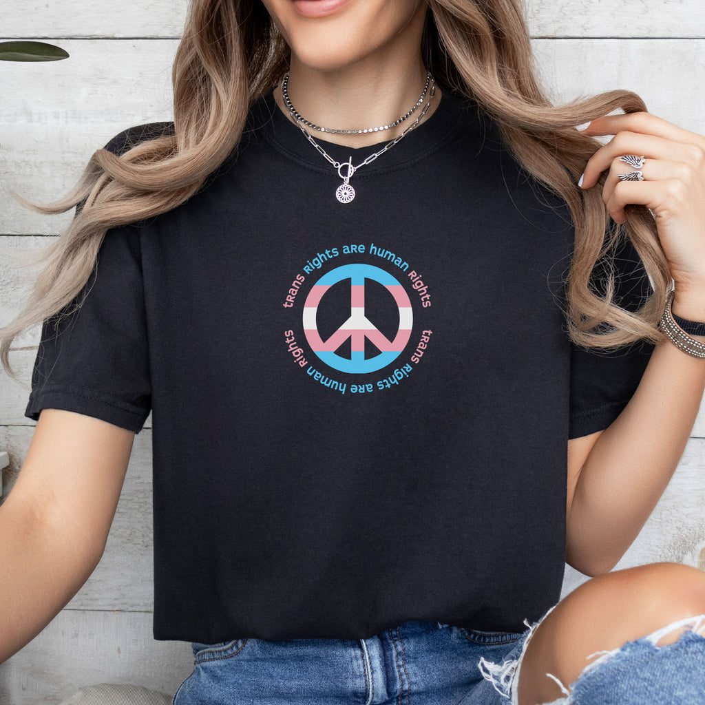 Female model wearing black t-shirt with a peace sign in transgender flag colors and the text “Trans Rights Are Human Rights” in a circular design.