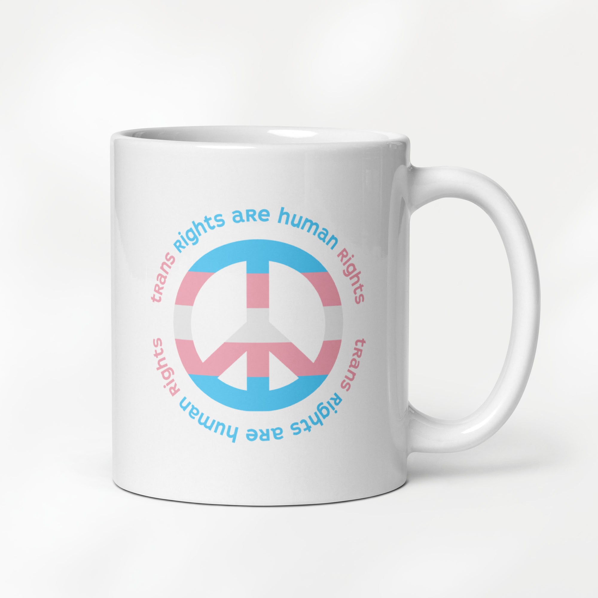 11 ounce ceramic mug with a peace sign in transgender flag colors and the text “Trans Rights Are Human Rights” in a circular design around the peace sign.