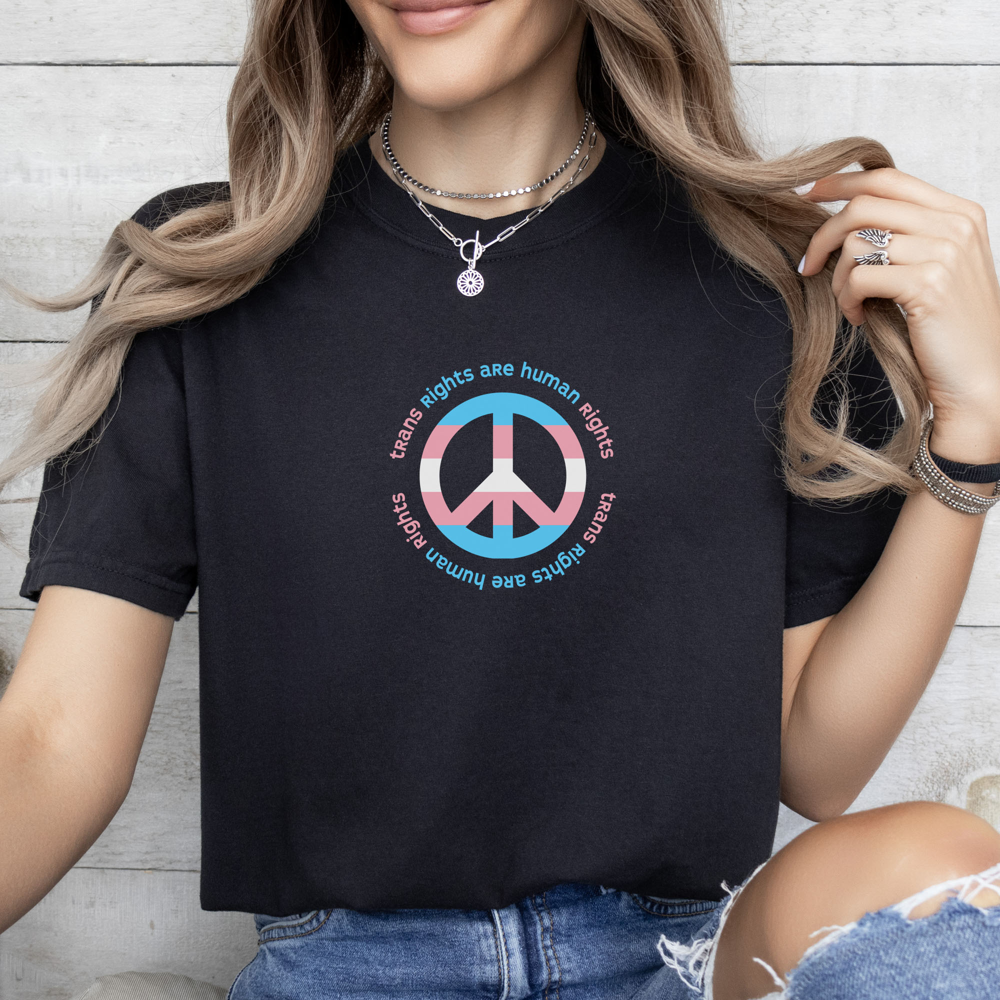 Female model wearing black t-shirt with a peace sign in transgender flag colors and the text “Trans Rights Are Human Rights” in a circular design.