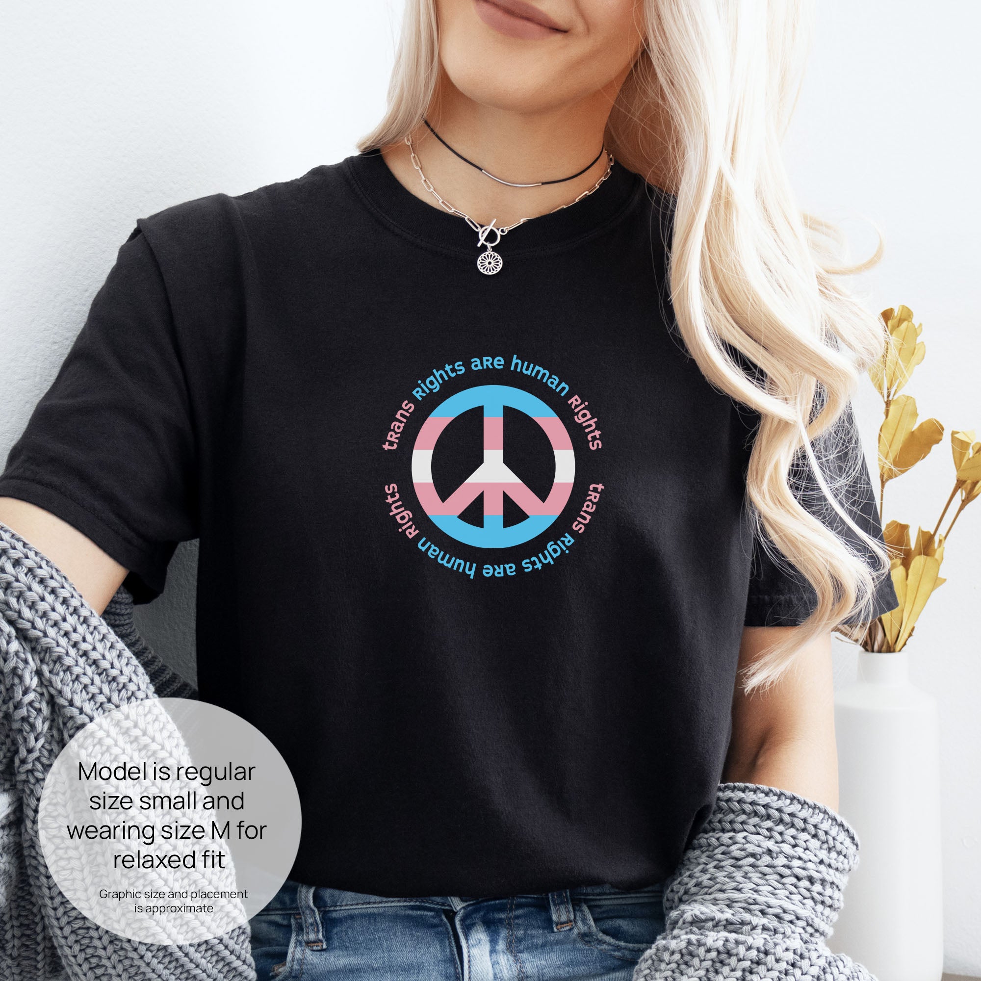 Female model wearing black t-shirt with a peace sign in transgender flag colors and the text “Trans Rights Are Human Rights” in a circular design. Includes size and fit information for the model who is regular size small, and wearing a size M shirt for a relaxed fit.
