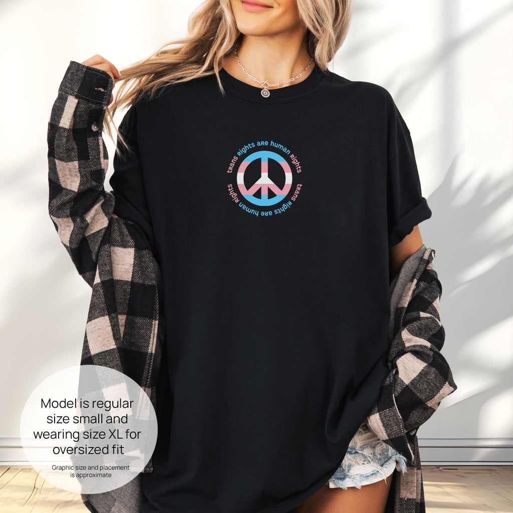 Female model wearing black t-shirt with a peace sign in transgender flag colors and the text “Trans Rights Are Human Rights” in a circular design. Includes size and fit information for the model who is regular size small, and wearing a size XL shirt for an oversized fit.
