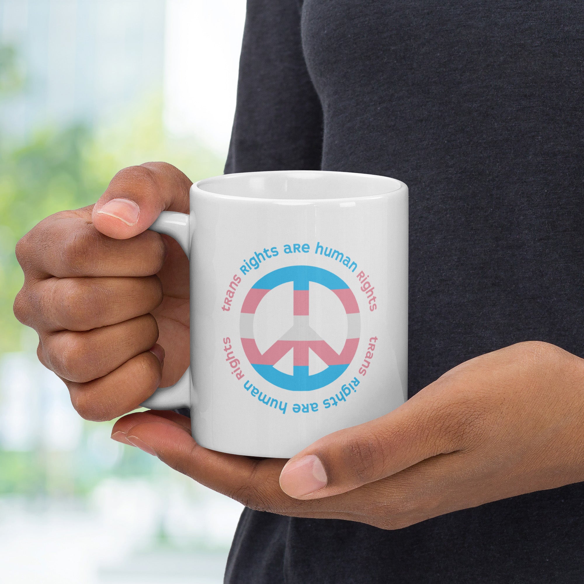 Person holding coffee mug with a peace sign in transgender flag colors and the text “Trans Rights Are Human Rights” in a circular design around the peace sign.
