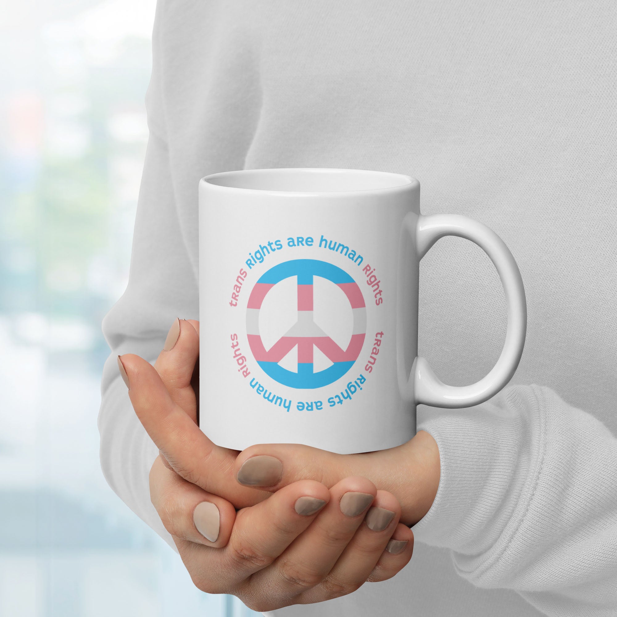 Person holding coffee mug with a peace sign in transgender flag colors and the text “Trans Rights Are Human Rights” in a circular design around the peace sign.