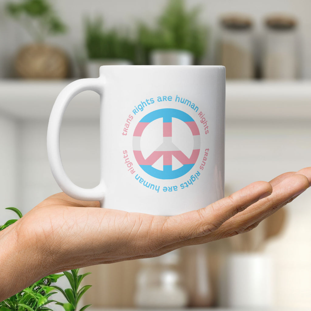 Coffee mug with a peace sign in transgender flag colors and the text “Trans Rights Are Human Rights” in a circular design around the peace sign, held in the palm of a hand.