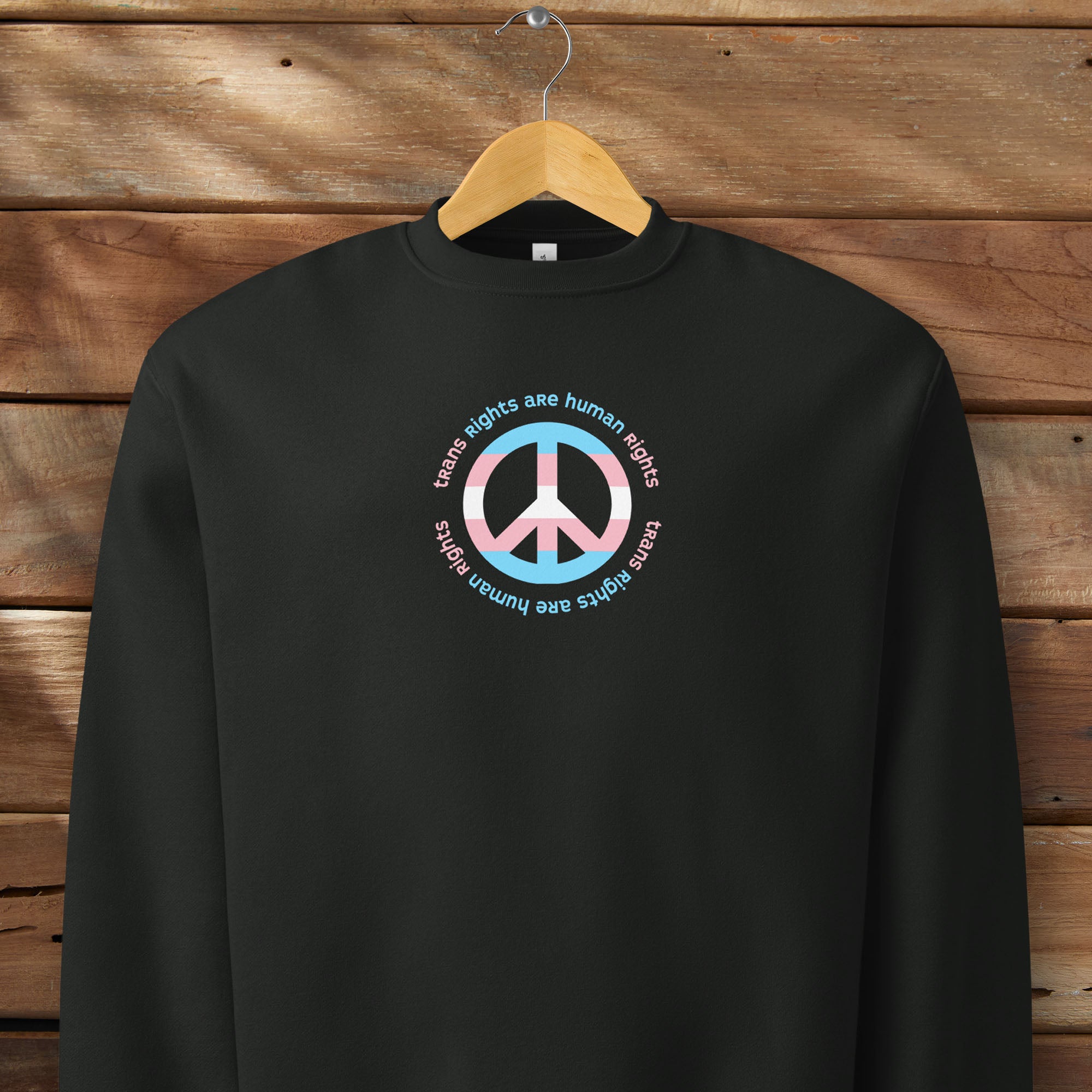 Black sweatshirt with a peace sign in transgender flag colors and the text “Trans Rights Are Human Rights” in a circular design around the peace sign. Hanging on a hanger.