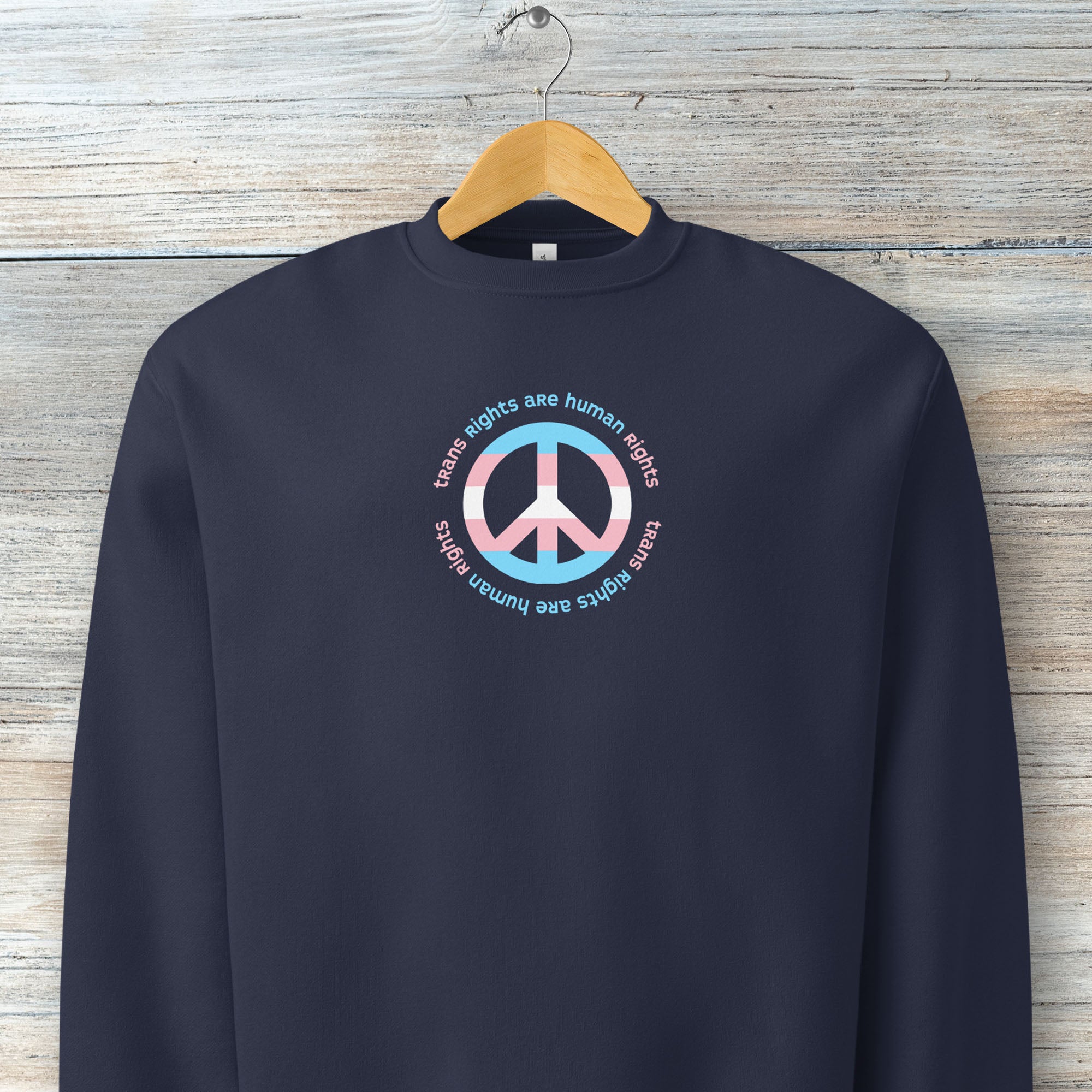 Navy sweatshirt with a peace sign in transgender flag colors and the text “Trans Rights Are Human Rights” in a circular design around the peace sign. Hanging on a hanger.
