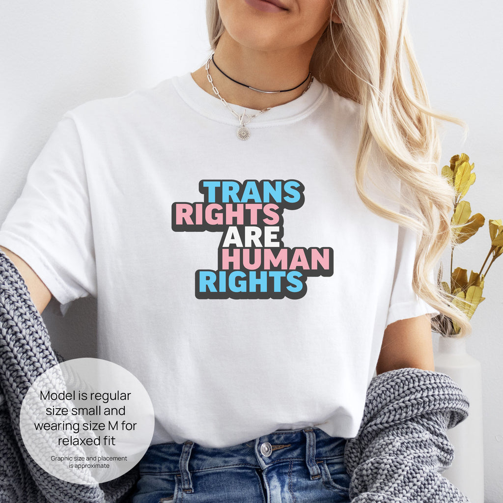 Female model wearing white t-shirt with graphic reading "trans rights are human rights" in the colors of the trans flag. Includes size and fit information for the model who is regular size small, and wearing a size M shirt for a relaxed fit.

