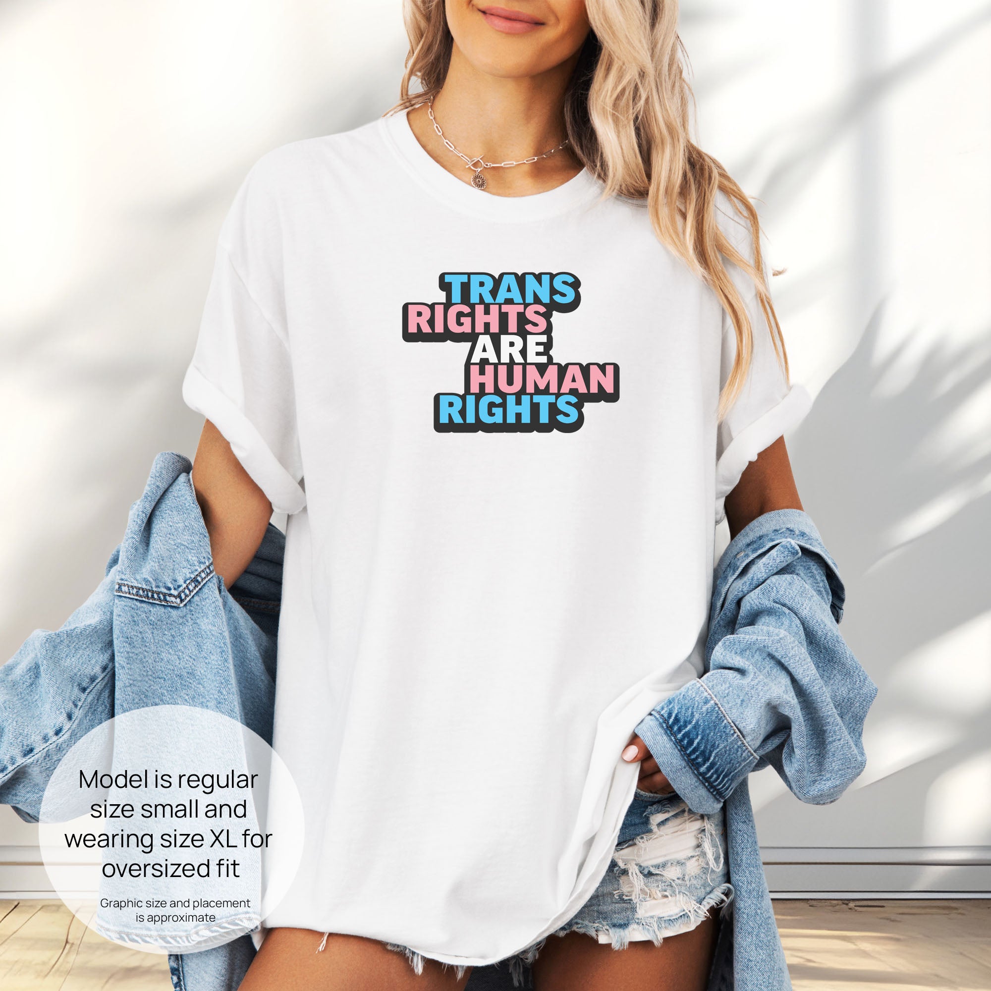 Female model wearing white t-shirt with graphic reading "trans rights are human rights" in the colors of the trans flag. Includes size and fit information for the model who is regular size small, and wearing a size XL shirt for an oversized fit.
