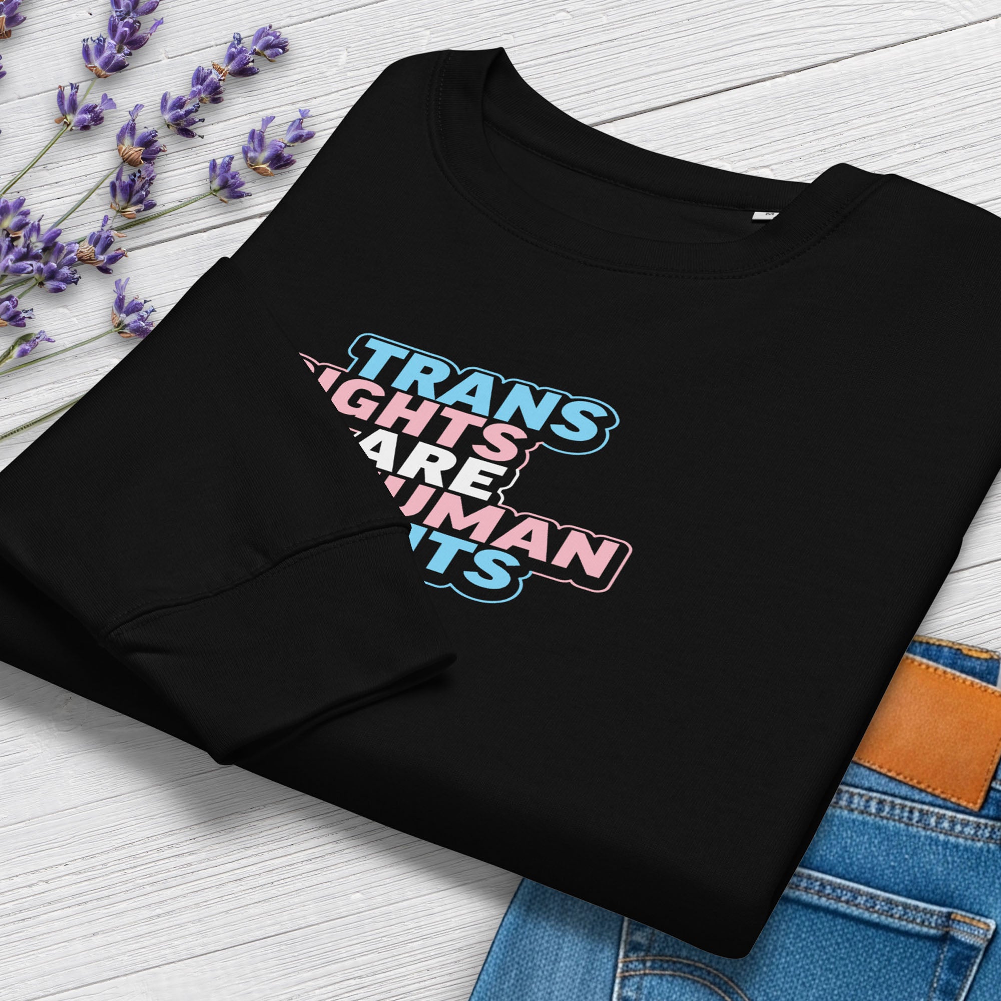 Folded black women's crewneck sweatshirt with graphic that reads "trans rights are human rights" in the light blue, pink, and white colors of the trans flag, showing cuff detail, styled with greenery and a pair of jeans.