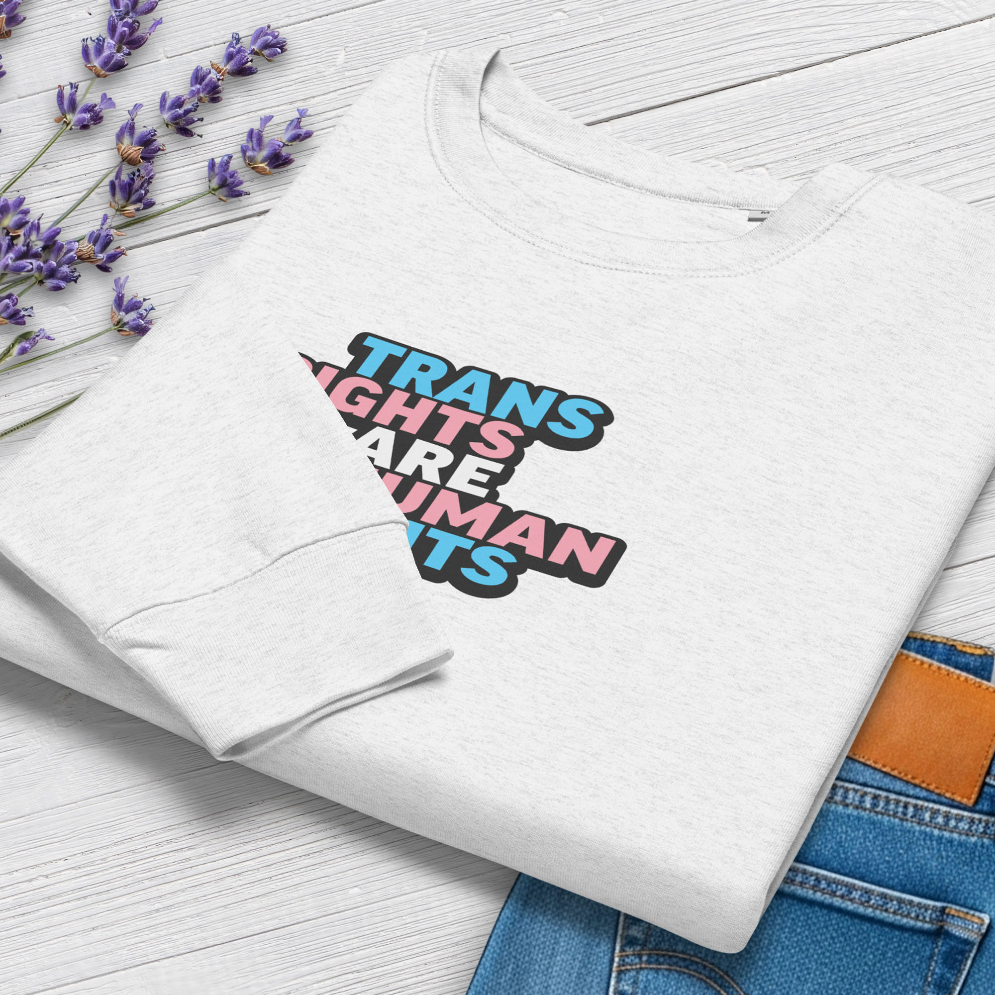 Folded grey women's crewneck sweatshirt with graphic that reads "trans rights are human rights" in the light blue, pink, and white colors of the trans flag, showing cuff detail, styled with greenery and a pair of jeans.