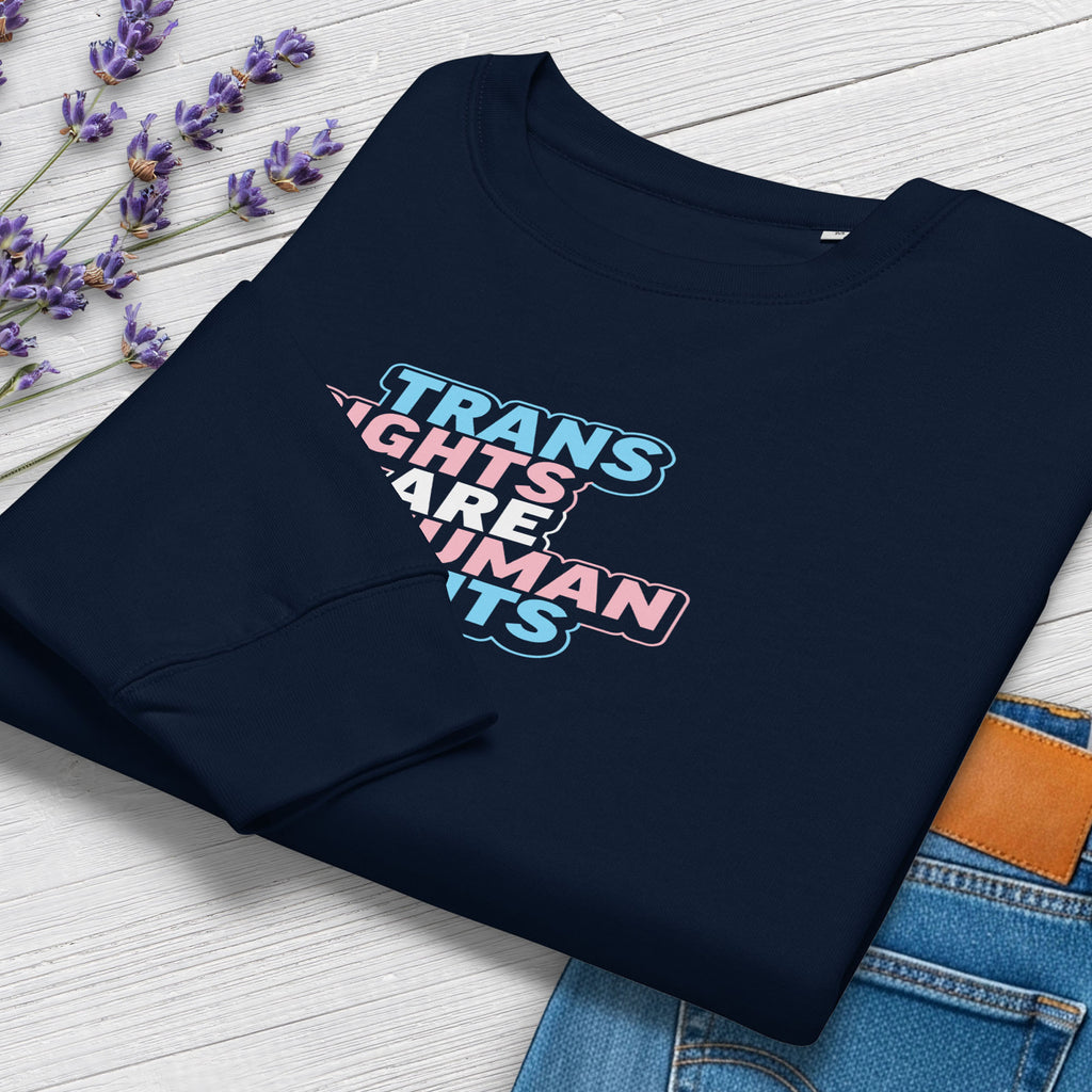 Folded navy women's crewneck sweatshirt with graphic that reads "trans rights are human rights" in the light blue, pink, and white colors of the trans flag, showing cuff detail, styled with greenery and a pair of jeans.