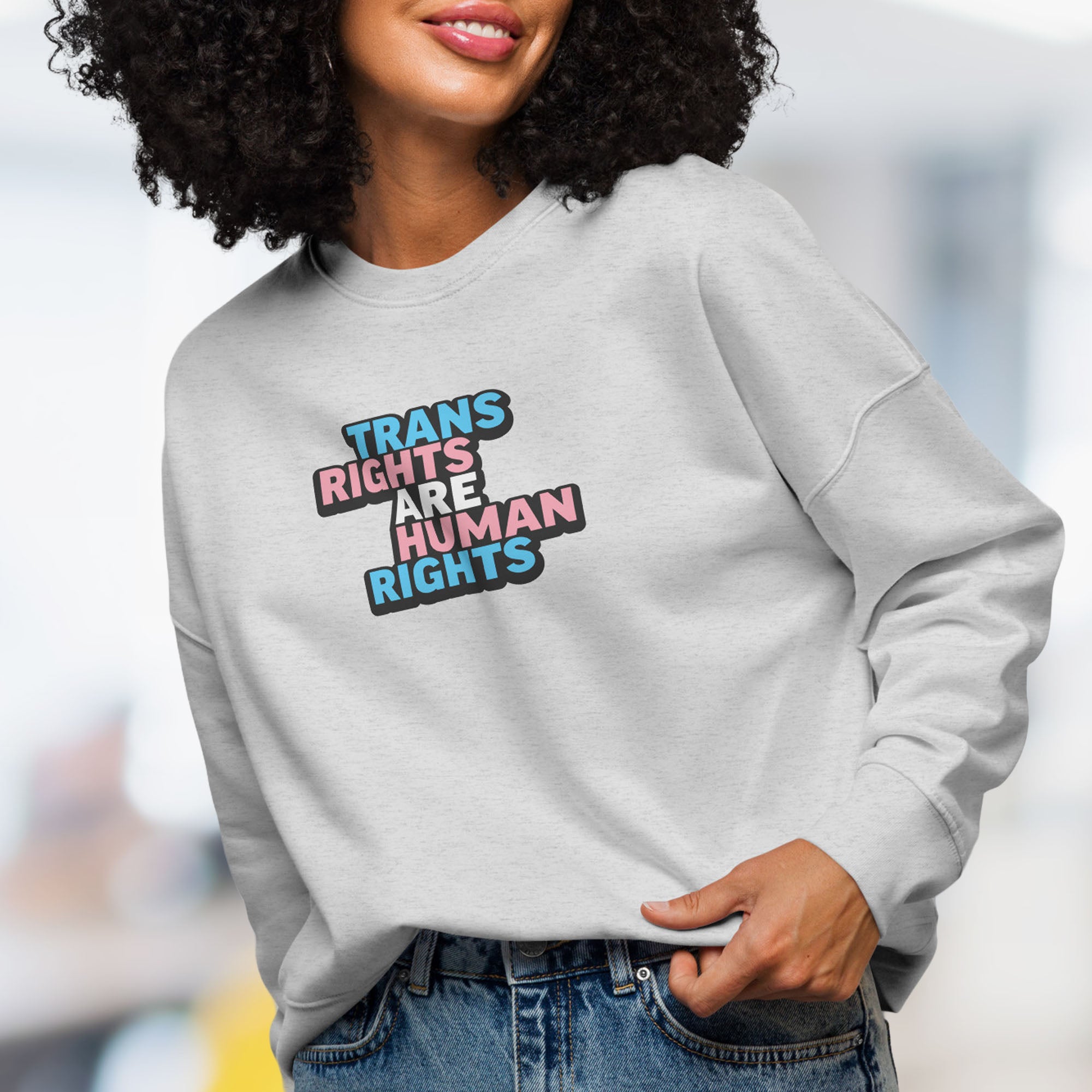 Female model wearing grey women's crewneck sweatshirt with graphic that reads "trans rights are human rights" in the light blue, pink, and white colors of the trans flag.