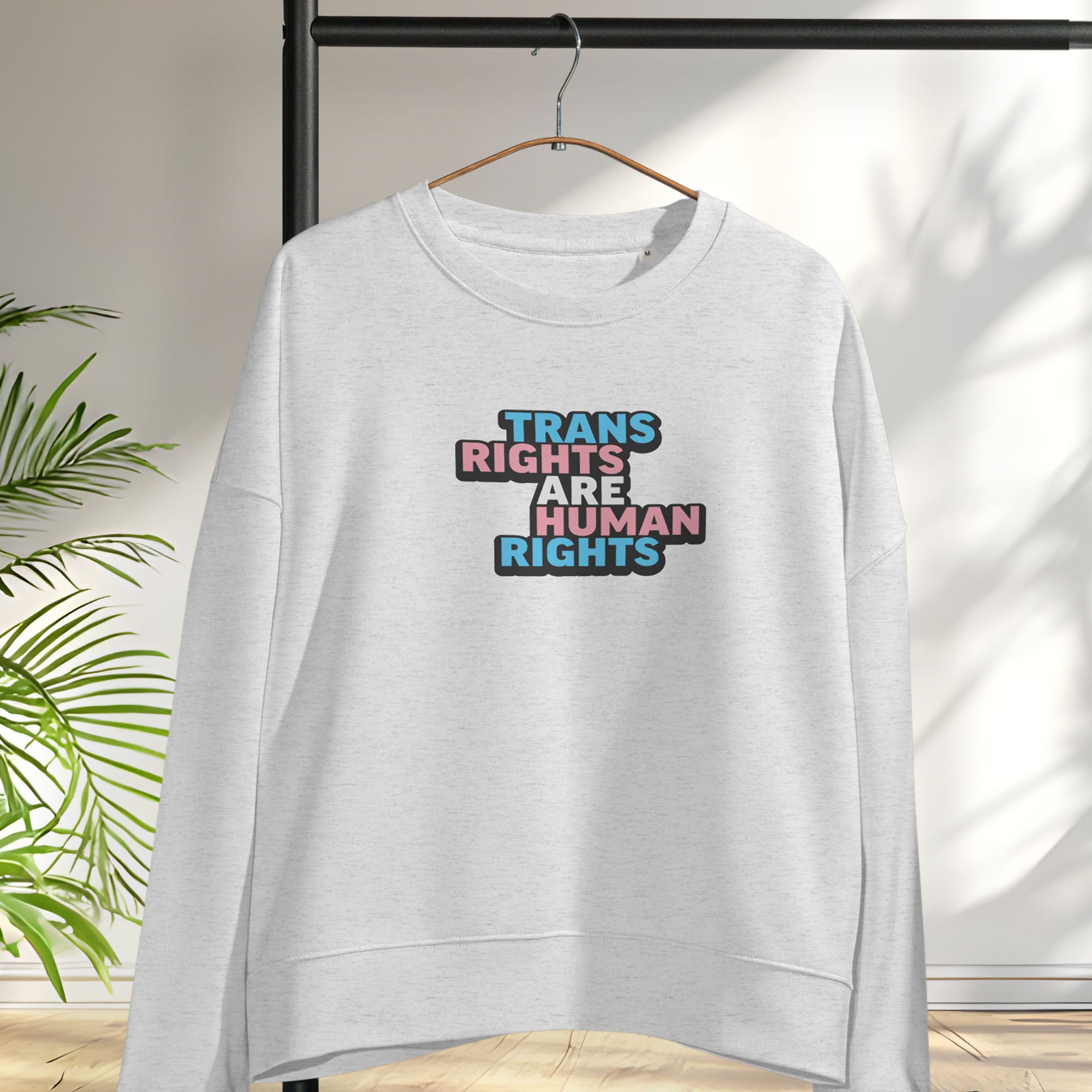 Hanging grey women's crewneck sweatshirt with graphic that reads "trans rights are human rights" in the light blue, pink, and white colors of the trans flag.