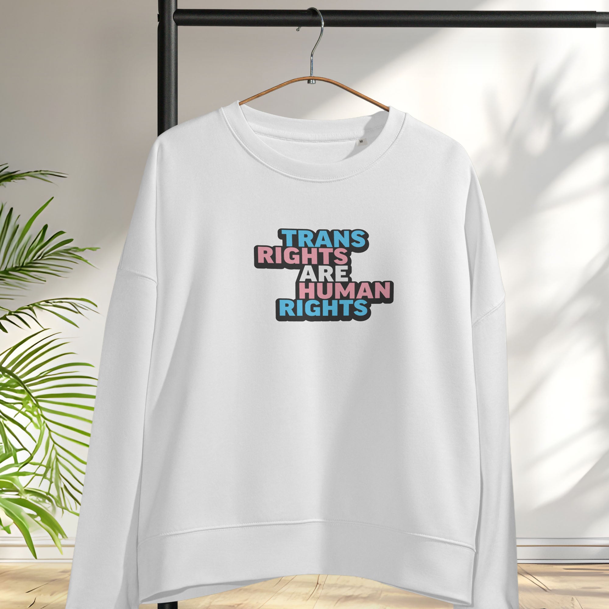 Hanging white women's crewneck sweatshirt with graphic that reads "trans rights are human rights" in the light blue, pink, and white colors of the trans flag.