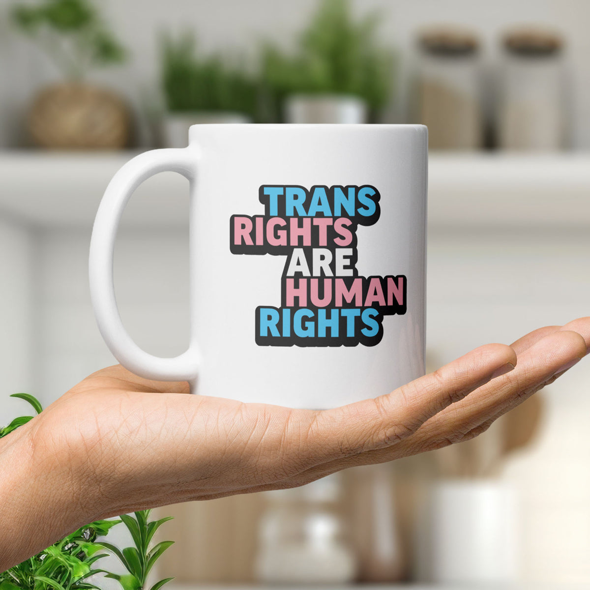 White ceramic mug with bold text “Trans Rights Are Human Rights” in blue, pink, and black colors representing the transgender pride flag, in the palm of a hand.