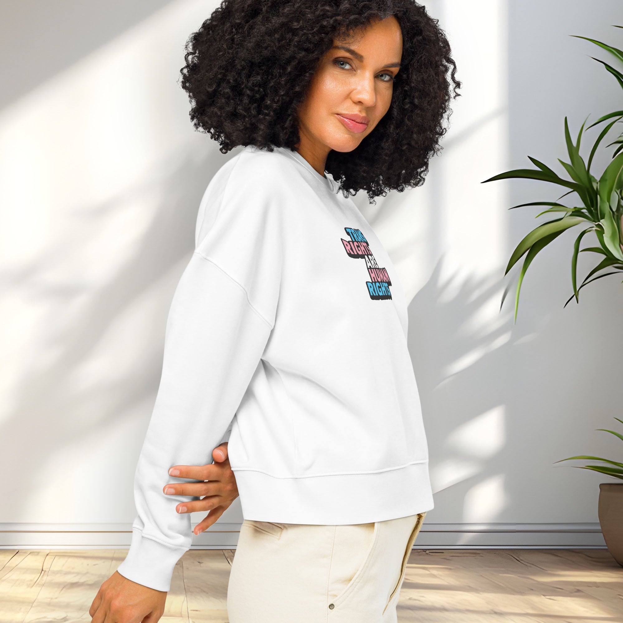 Side view of female model wearing white women's crewneck sweatshirt with graphic that reads "trans rights are human rights" in the light blue, pink, and white colors of the trans flag, showing shirt fit.