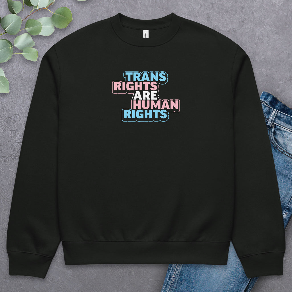 Black crewneck sweatshirt with graphic that reads "trans rights are human rights" in the light blue, pink, and white colors of the trans flag, with a colorful outline around the text block. Laying flat with a pair of jeans.