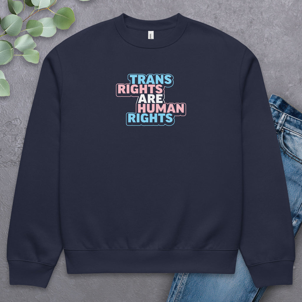 Black crewneck sweatshirt with graphic that reads "trans rights are human rights" in the light blue, pink, and white colors of the trans flag, with a colorful outline around the text block. Laying flat with a pair of jeans.