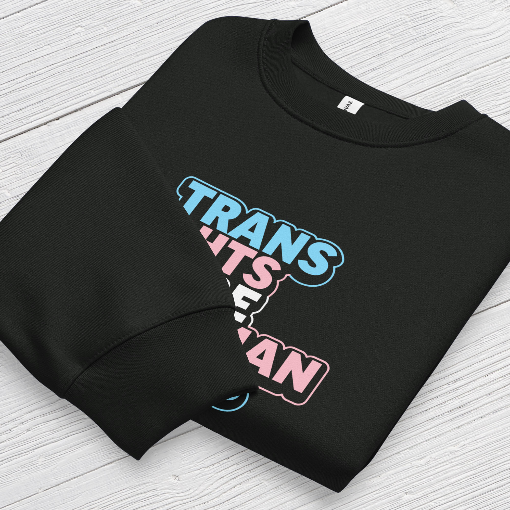 Folded black crewneck sweatshirt with graphic that reads "trans rights are human rights" in the light blue, pink, and white colors of the trans flag, with a colorful outline around the text block.