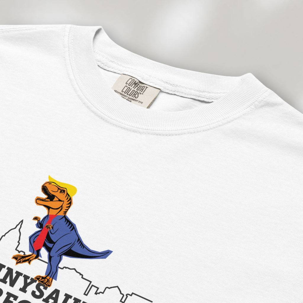 Closeup detail of white t-shirt with a funny drawing of an orange t-rex dinosaur dressed in a blue suit with a red tie that's too long, stupid yellow hair, and tiny hands. The dinosaur is stepping on a line drawing of the Washington DC skyline. Under the dinosaur is bold text that reads "tyranny saurus wrecks."