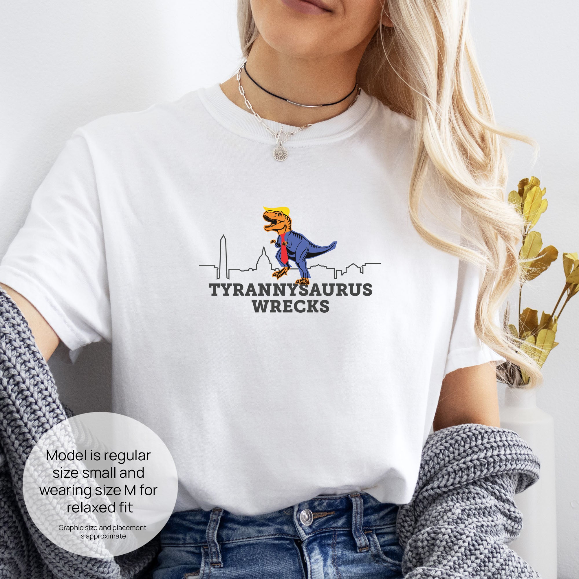Female model wearing white t-shirt with a funny drawing of an orange t-rex dinosaur. Includes size and fit information for the model who is regular size small, and wearing a size M shirt for a relaxed fit. T-Rex is dressed in a blue suit with a red tie that's too long, stupid yellow hair, and tiny hands. The dinosaur is stepping on a line drawing of the Washington DC skyline. Under the dinosaur is bold text that reads "tyranny saurus wrecks."