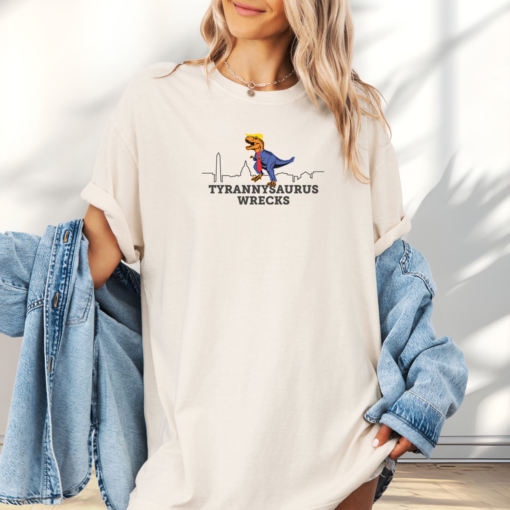 Female model wearing oversized ivory t-shirt with a funny drawing of an orange t-rex dinosaur dressed in a blue suit with a red tie that's too long, stupid yellow hair, and tiny hands. The dinosaur is stepping on a line drawing of the Washington DC skyline. Under the dinosaur is bold text that reads "tyranny saurus wrecks."