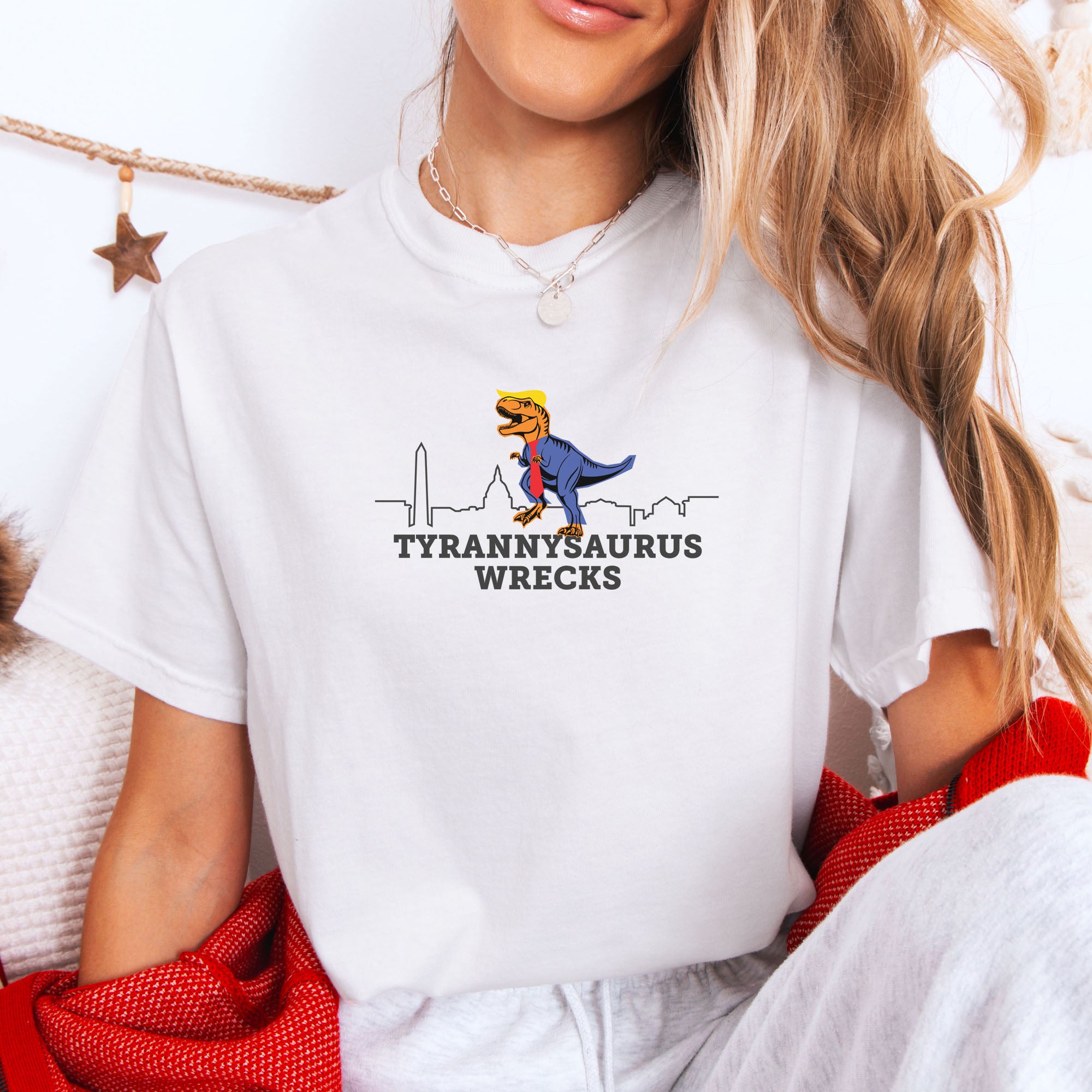 Female model wearing white t-shirt with a funny drawing of an orange t-rex dinosaur dressed in a blue suit with a red tie that's too long, stupid yellow hair, and tiny hands. The dinosaur is stepping on a line drawing of the Washington DC skyline. Under the dinosaur is bold text that reads "tyranny saurus wrecks."