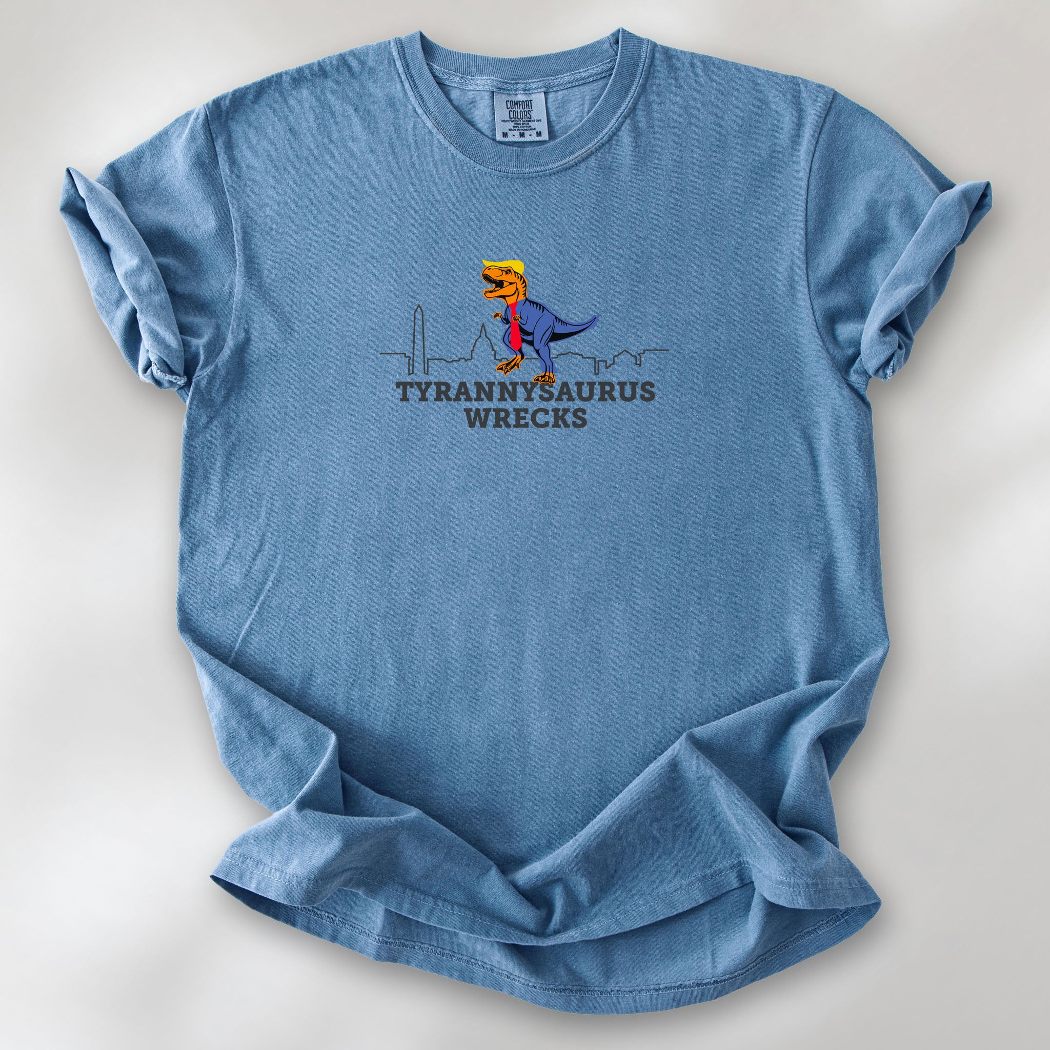 Blue jean t-shirt with a funny drawing of an orange t-rex dinosaur dressed in a blue suit with a red tie that's too long, stupid yellow hair, and tiny hands. The dinosaur is stepping on a line drawing of the Washington DC skyline. Under the dinosaur is bold text that reads "tyranny saurus wrecks."