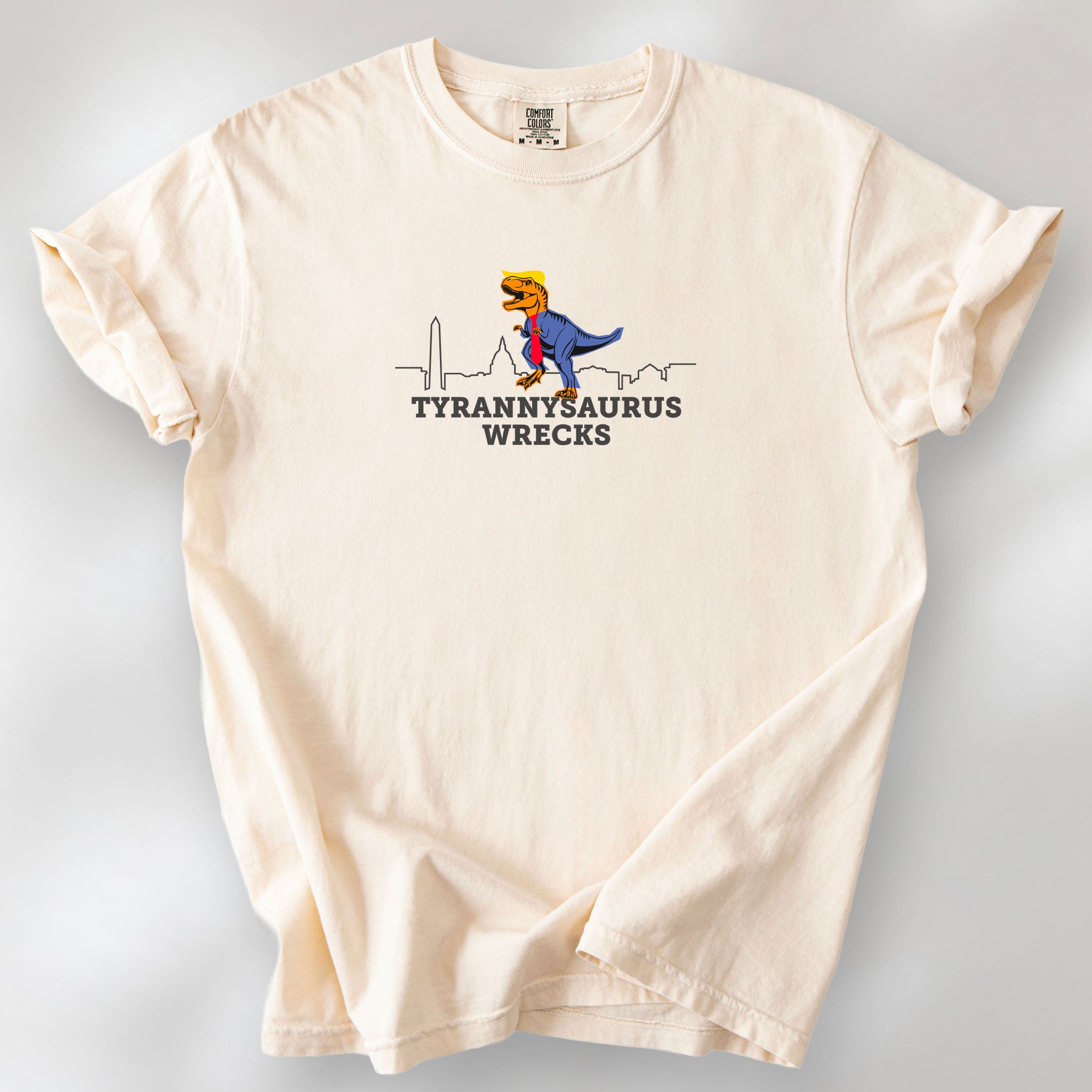 Ivory t-shirt with a funny drawing of an orange t-rex dinosaur dressed in a blue suit with a red tie that's too long, stupid yellow hair, and tiny hands. The dinosaur is stepping on a line drawing of the Washington DC skyline. Under the dinosaur is bold text that reads "tyranny saurus wrecks."