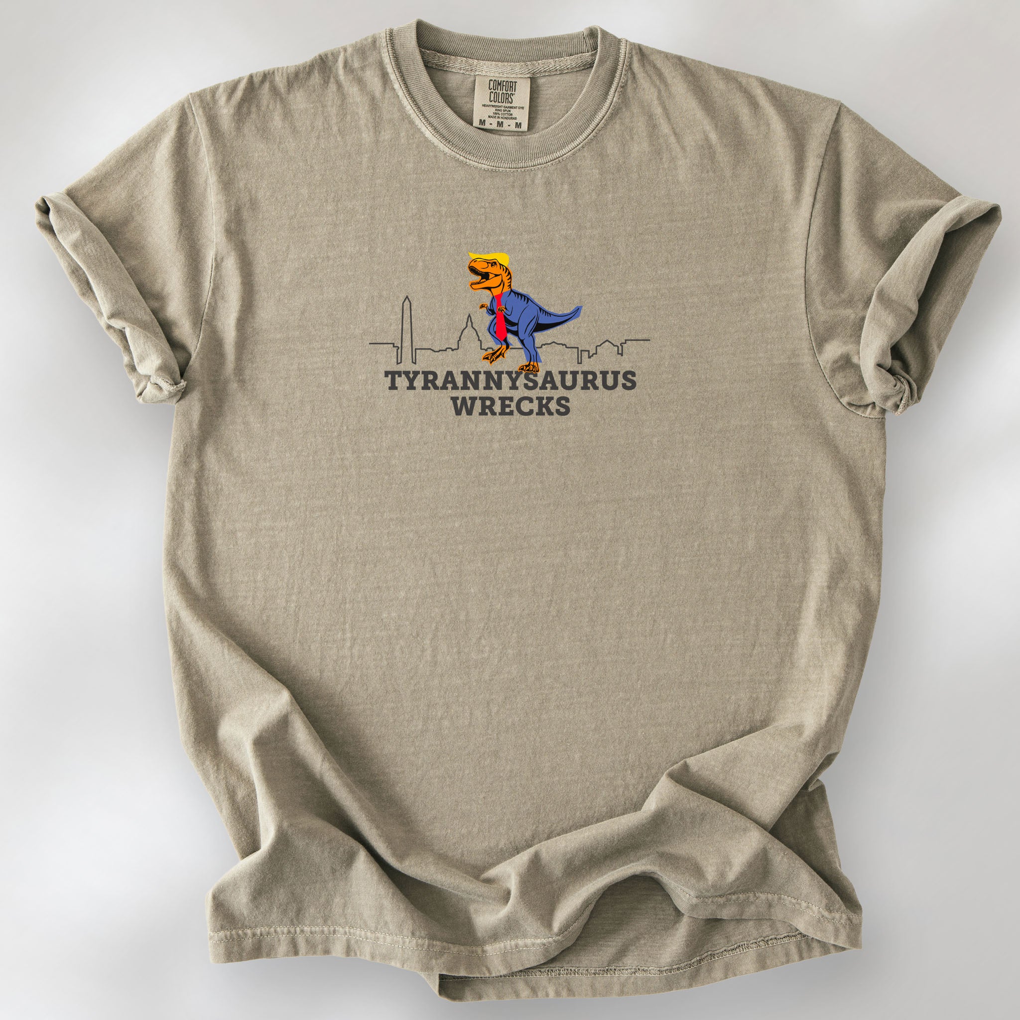 Khaki t-shirt with a funny drawing of an orange t-rex dinosaur. Includes size and fit information for the model who is 6 feet 0 inches, 176 pounds, and wearing a size XL shirt. T-Rex is dressed in a blue suit with a red tie that's too long, stupid yellow hair, and tiny hands. The dinosaur is stepping on a line drawing of the Washington DC skyline. Under the dinosaur is bold text that reads "tyranny saurus wrecks."