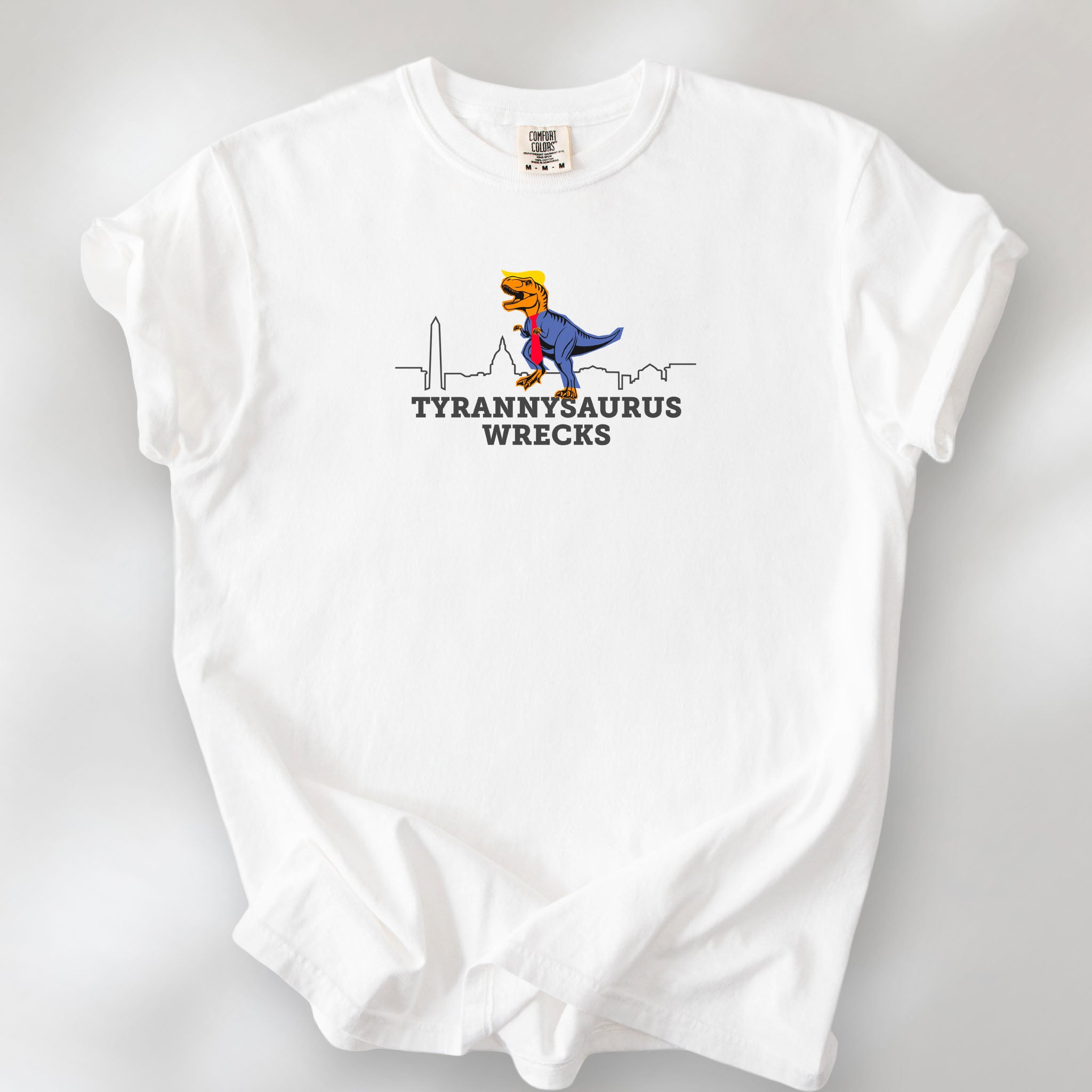 White t-shirt with a funny drawing of an orange t-rex dinosaur dressed in a blue suit with a red tie that's too long, stupid yellow hair, and tiny hands. The dinosaur is stepping on a line drawing of the Washington DC skyline. Under the dinosaur is bold text that reads "tyranny saurus wrecks."