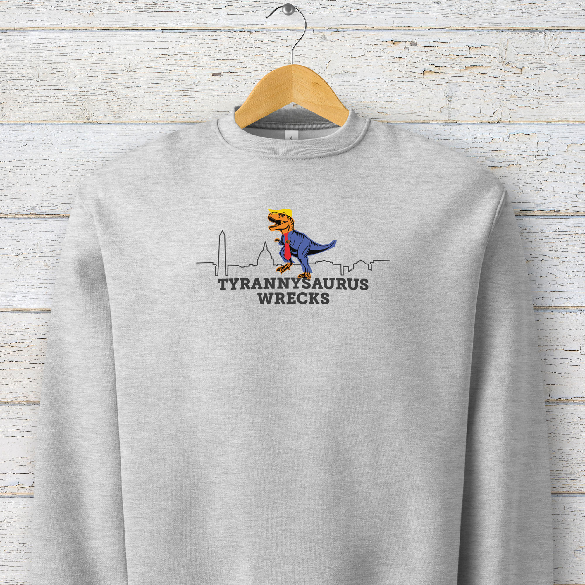 Athletic heather grey sweatshirt with a funny drawing of an orange t-rex dinosaur dressed in a blue suit with a red tie that's too long, stupid yellow hair, and tiny hands. The dinosaur is stepping on a line drawing of the Washington DC skyline. Under the dinosaur is bold text that reads "tyranny saurus wrecks." It's hanging on a hanger.