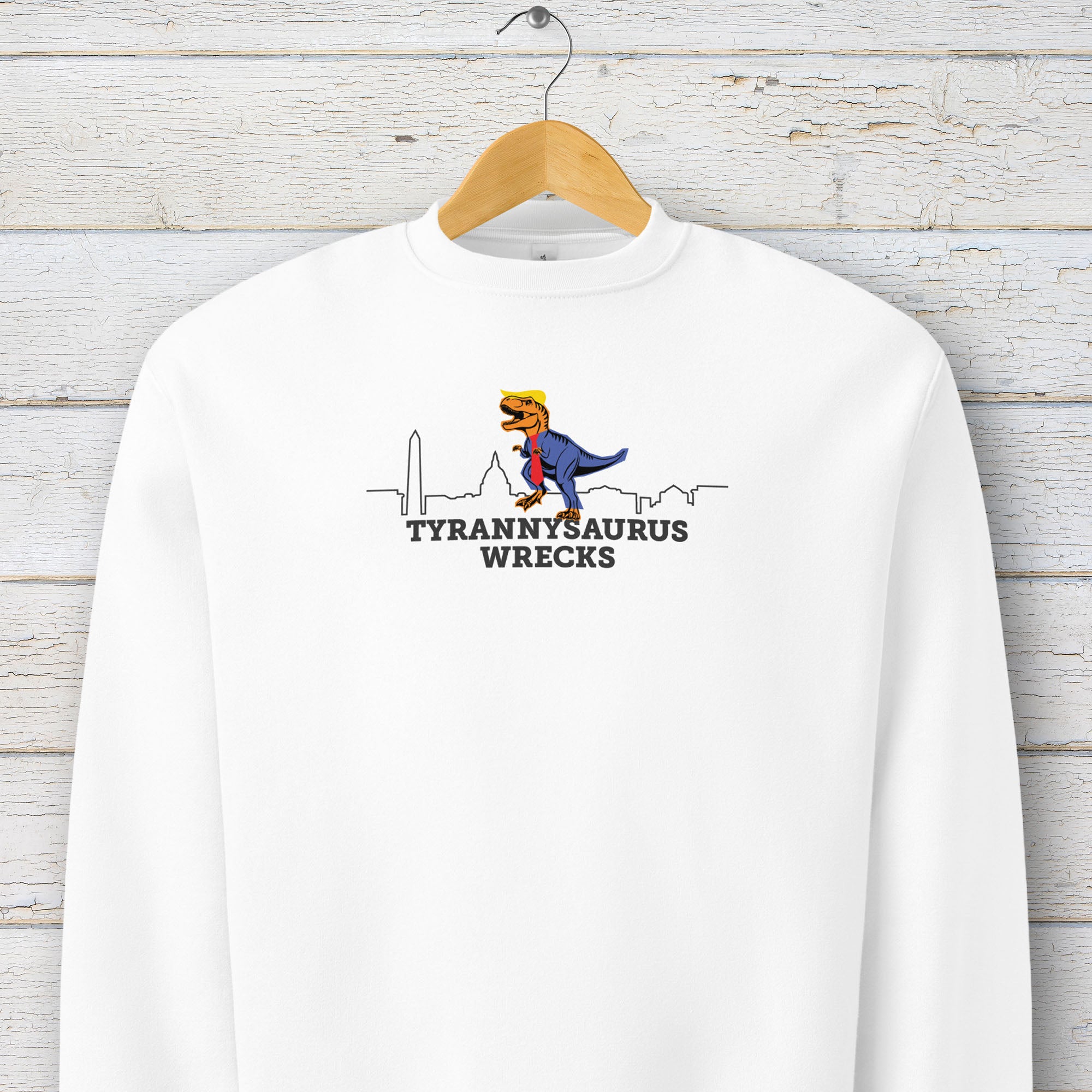 White sweatshirt with a funny drawing of an orange t-rex dinosaur dressed in a blue suit with a red tie that's too long, stupid yellow hair, and tiny hands. The dinosaur is stepping on a line drawing of the Washington DC skyline. Under the dinosaur is bold text that reads "tyranny saurus wrecks." It's hanging on a hanger.